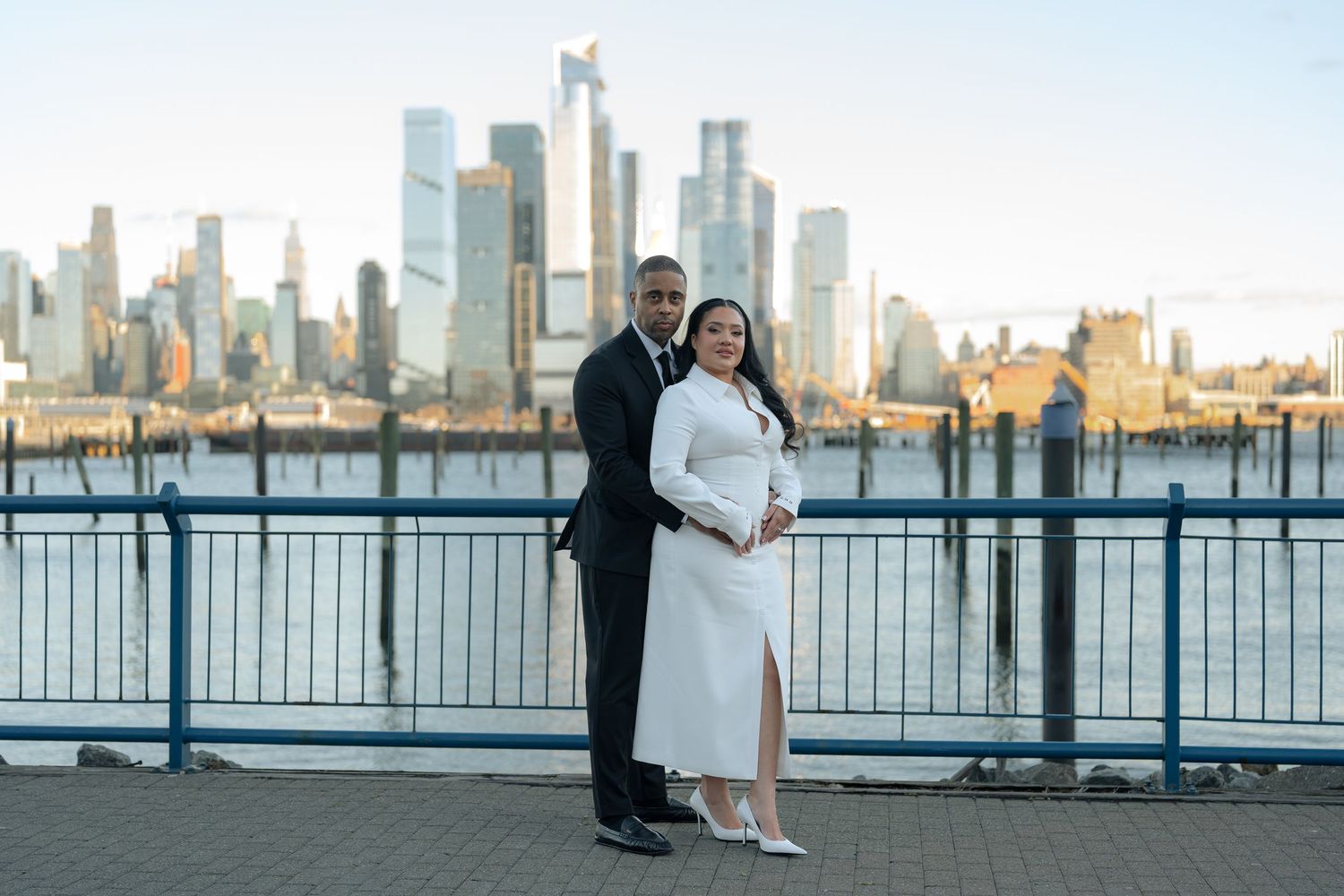Couple posing along waterfront railing with city skyline visible in background at sunset.
