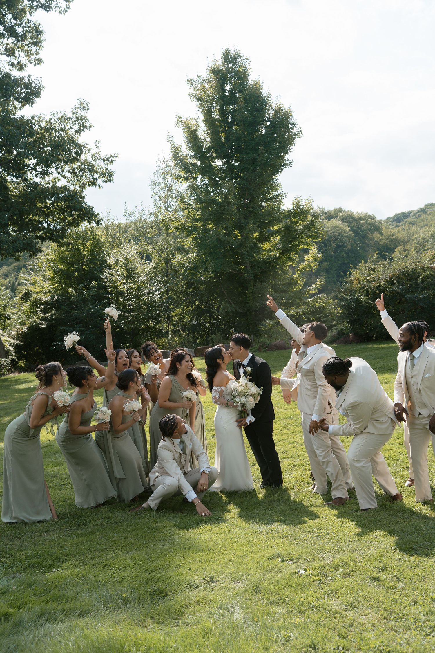 Wedding party celebrates on green lawn with bridesmaids in sage gowns and groomsmen in cream suits as couple shares a kiss.
