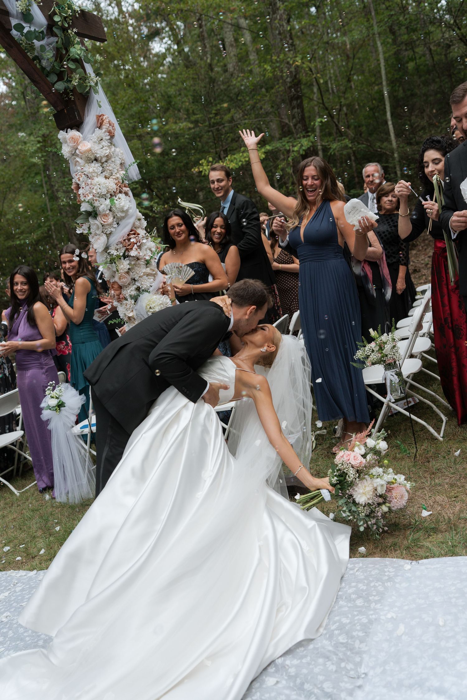 Groom dips bride for a kiss at outdoor wedding ceremony while guests cheer and blow bubbles.