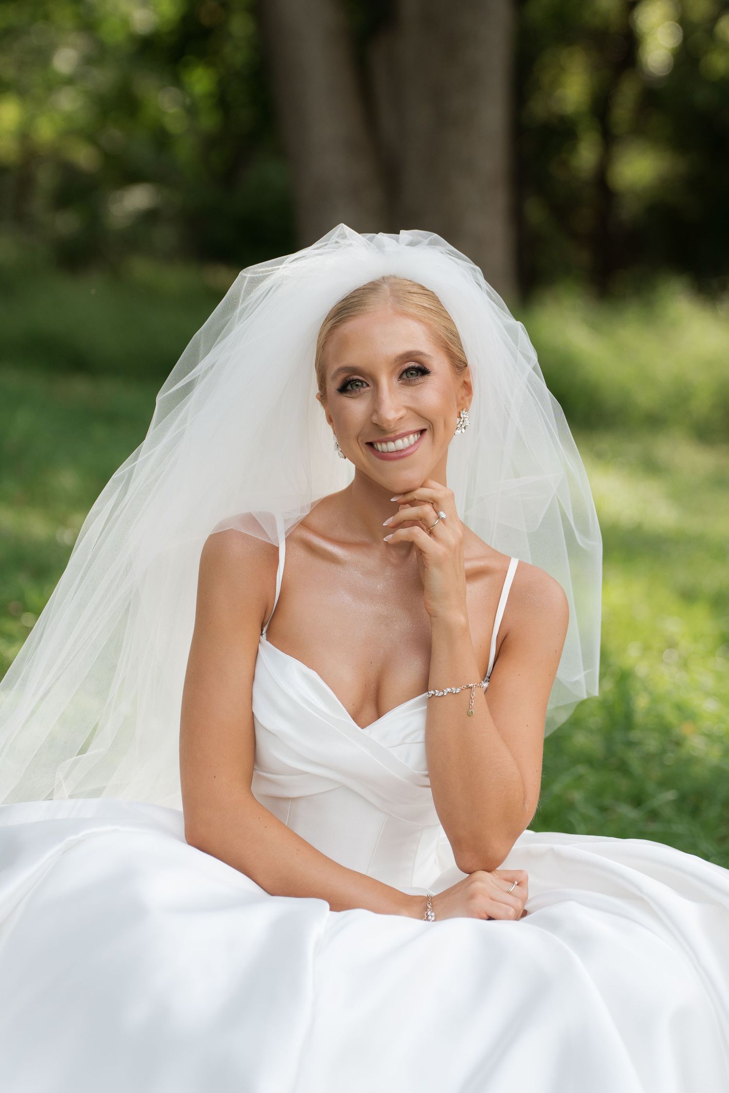 Smiling bride in white wedding gown and veil sitting outdoors in lush green garden setting.