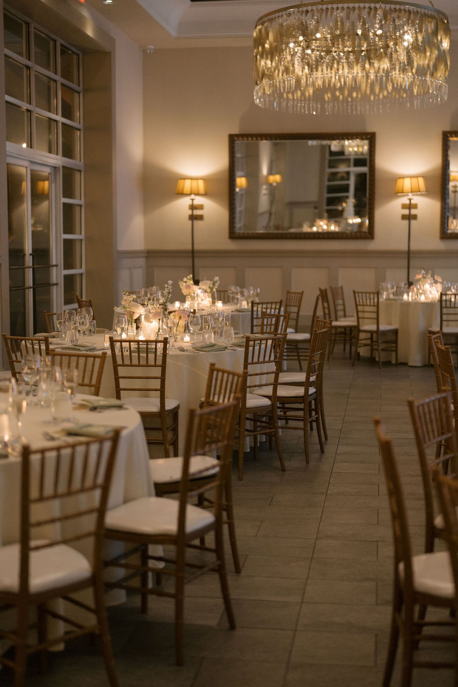 Elegant wedding reception hall with gold chiavari chairs, white tablecloths, candles, and a crystal chandelier.
