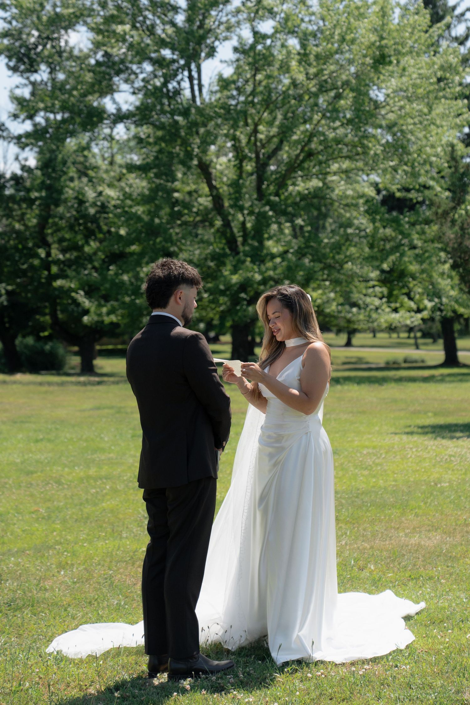 Bride placing wedding ring on groom's finger during outdoor ceremony in lush green park.