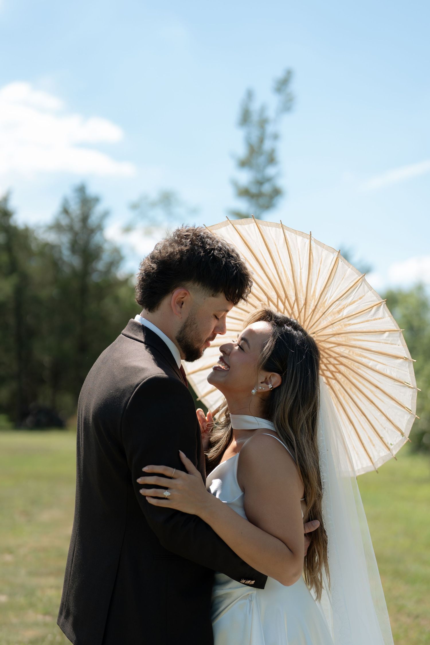 Wedding couple nearly kissing under white parasol umbrella in sunny outdoor field on wedding day.