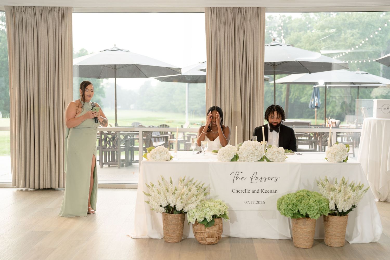 Bride and groom seated at wedding reception sweetheart table while bridesmaid delivers a heartfelt toast.