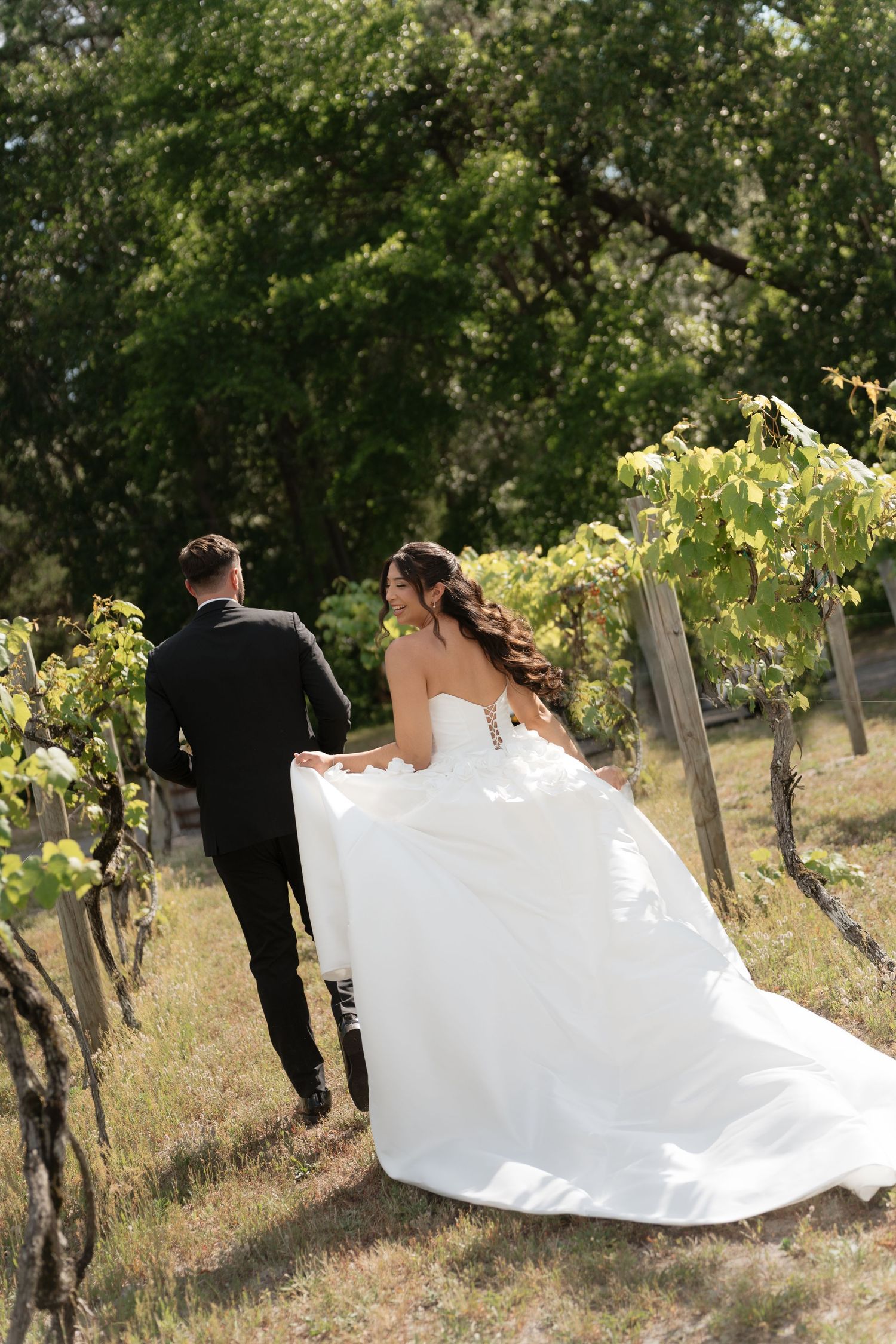 Bride in flowing white gown and groom in black suit walking hand in hand through sunny vineyard.