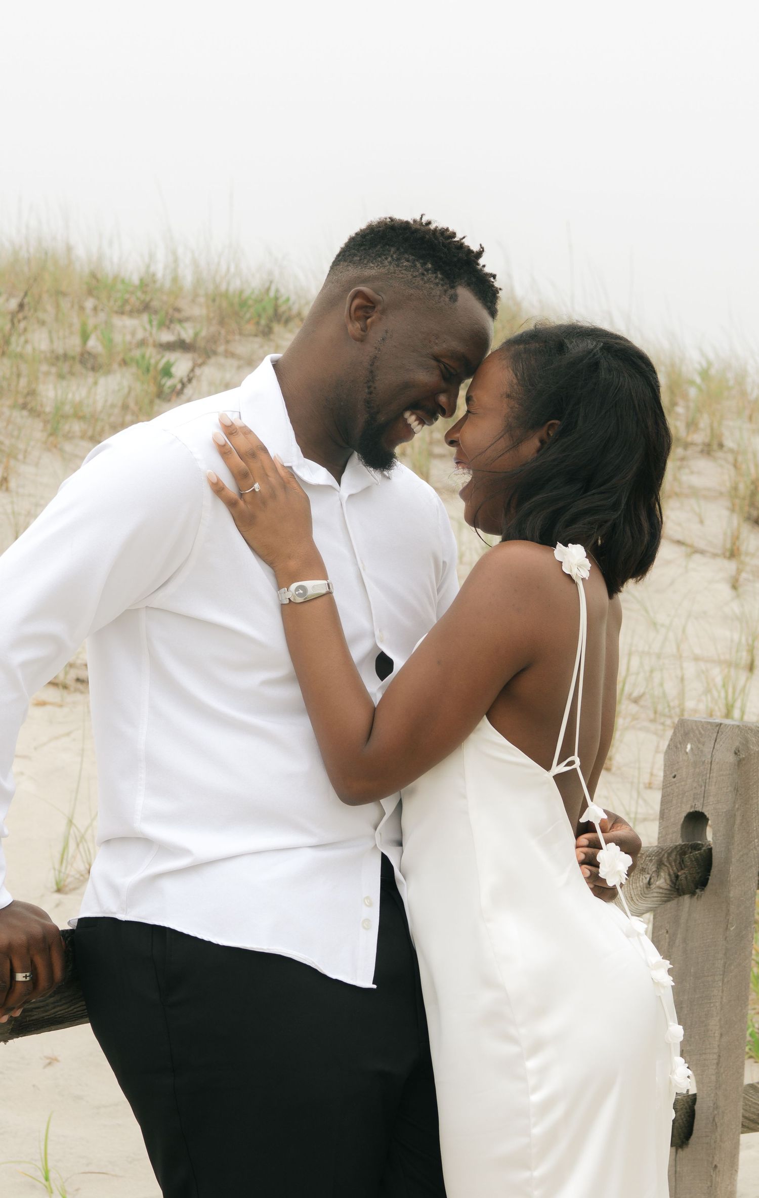A romantic couple shares an intimate moment on a sandy beach, with the woman wearing a white dress and man in white shirt.