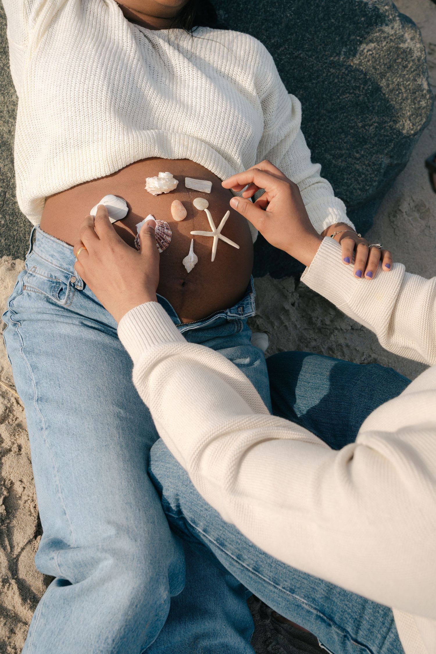 Person wearing light blue jeans and white sweater sits on textured stone surface in casual pose.