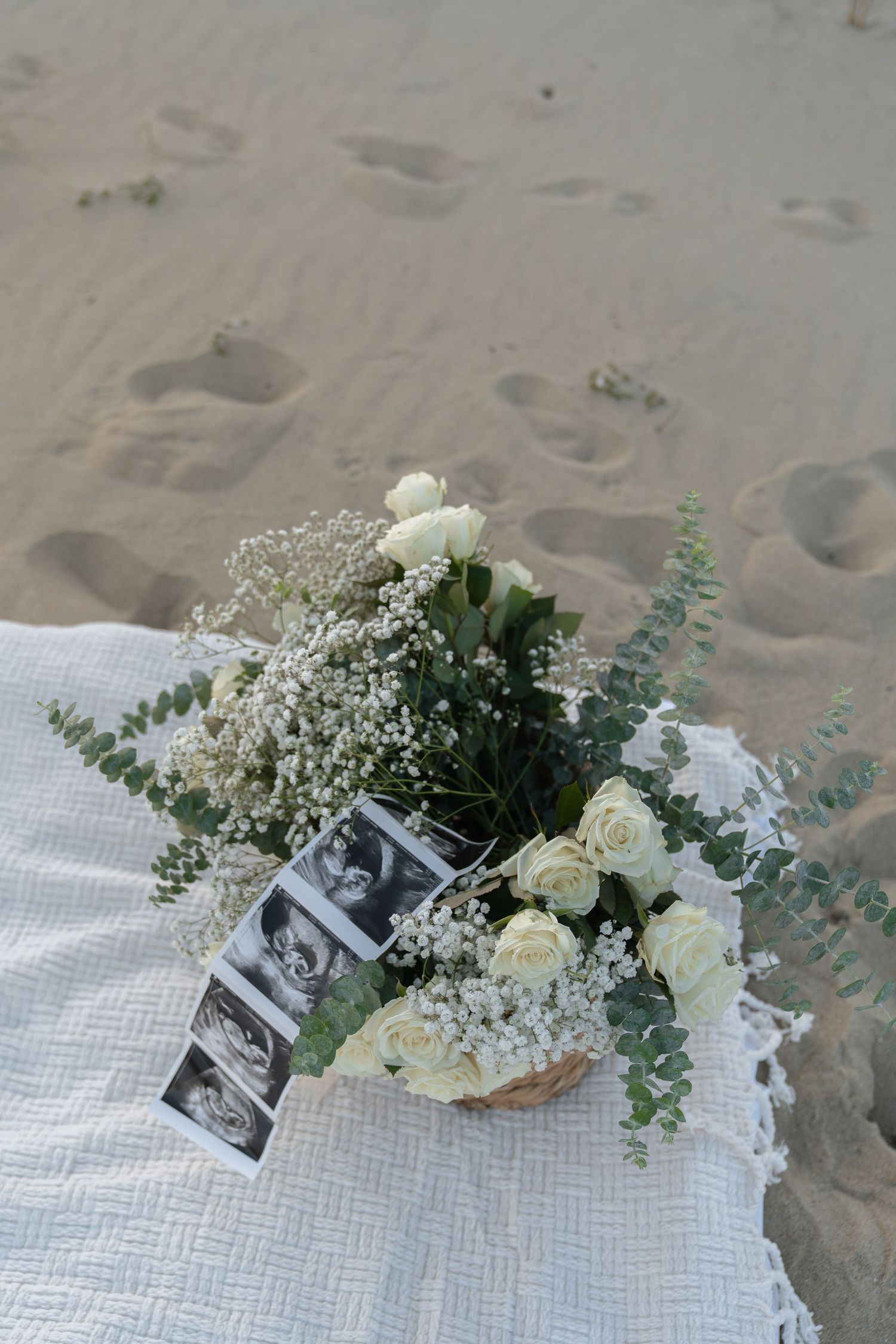 White roses and baby's breath bouquet with ultrasound photos on white blanket in sand at beach maternity shoot.