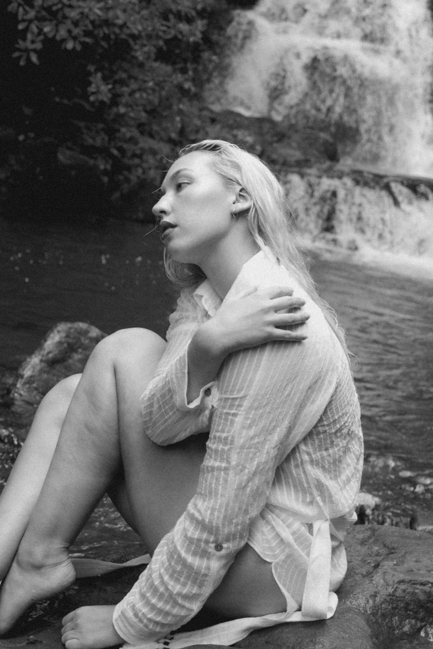 Artistic black and white portrait by a waterfall shows figure in white clothing reclining on rocks.