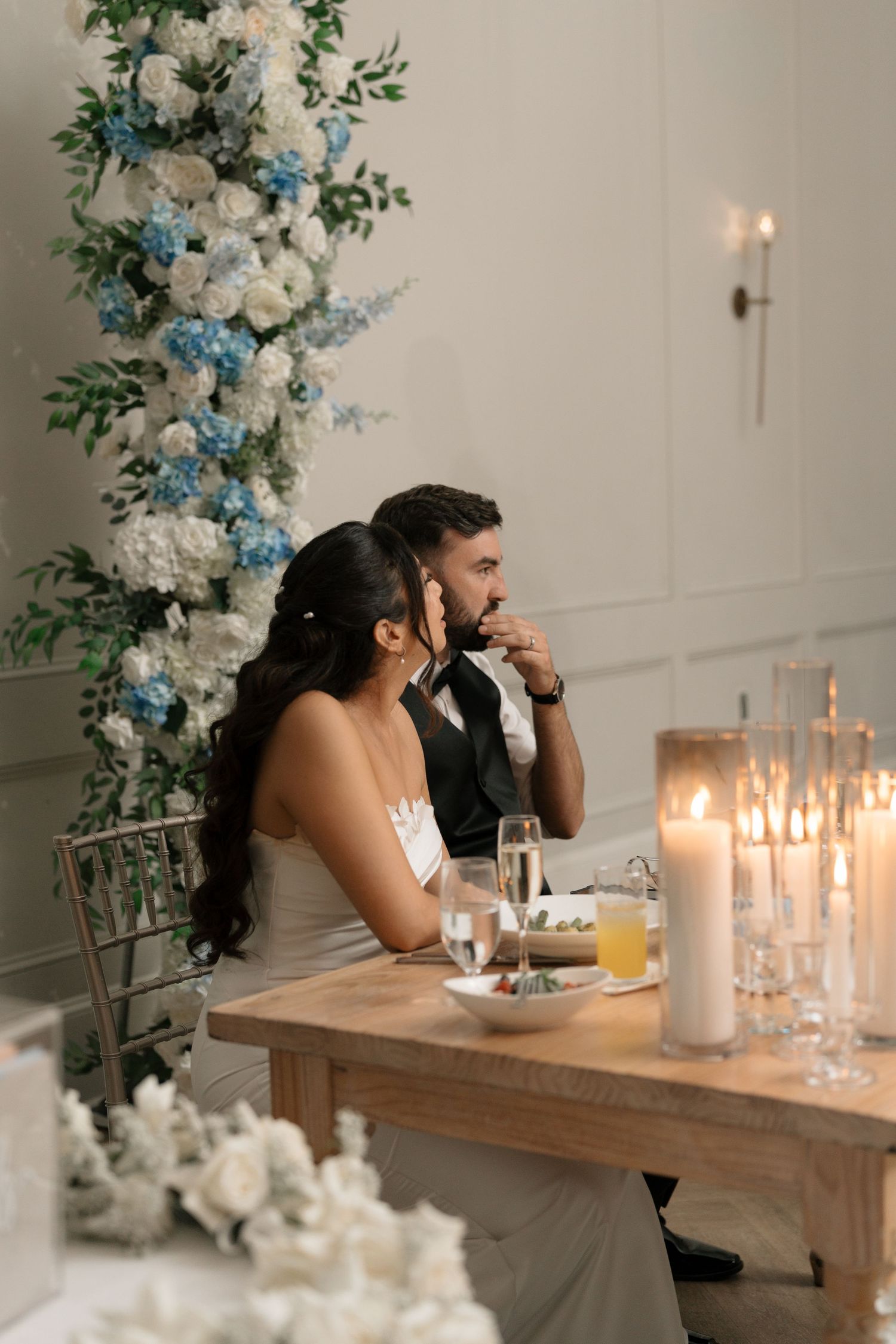 Bride and groom sit at sweetheart table adorned with blue and white floral arrangement.
