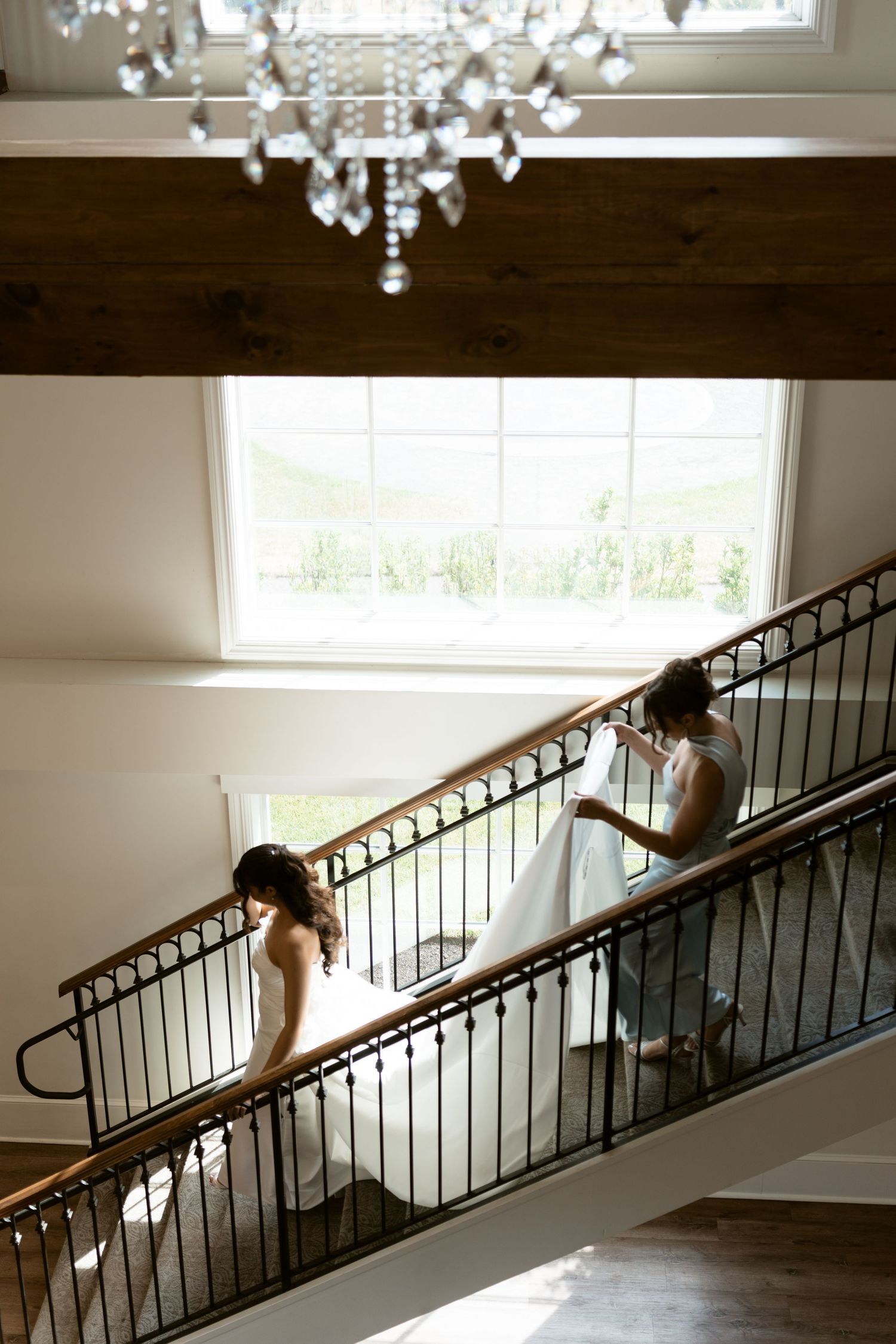 Bride in white gown walking down elegant staircase with crystal chandelier overhead.