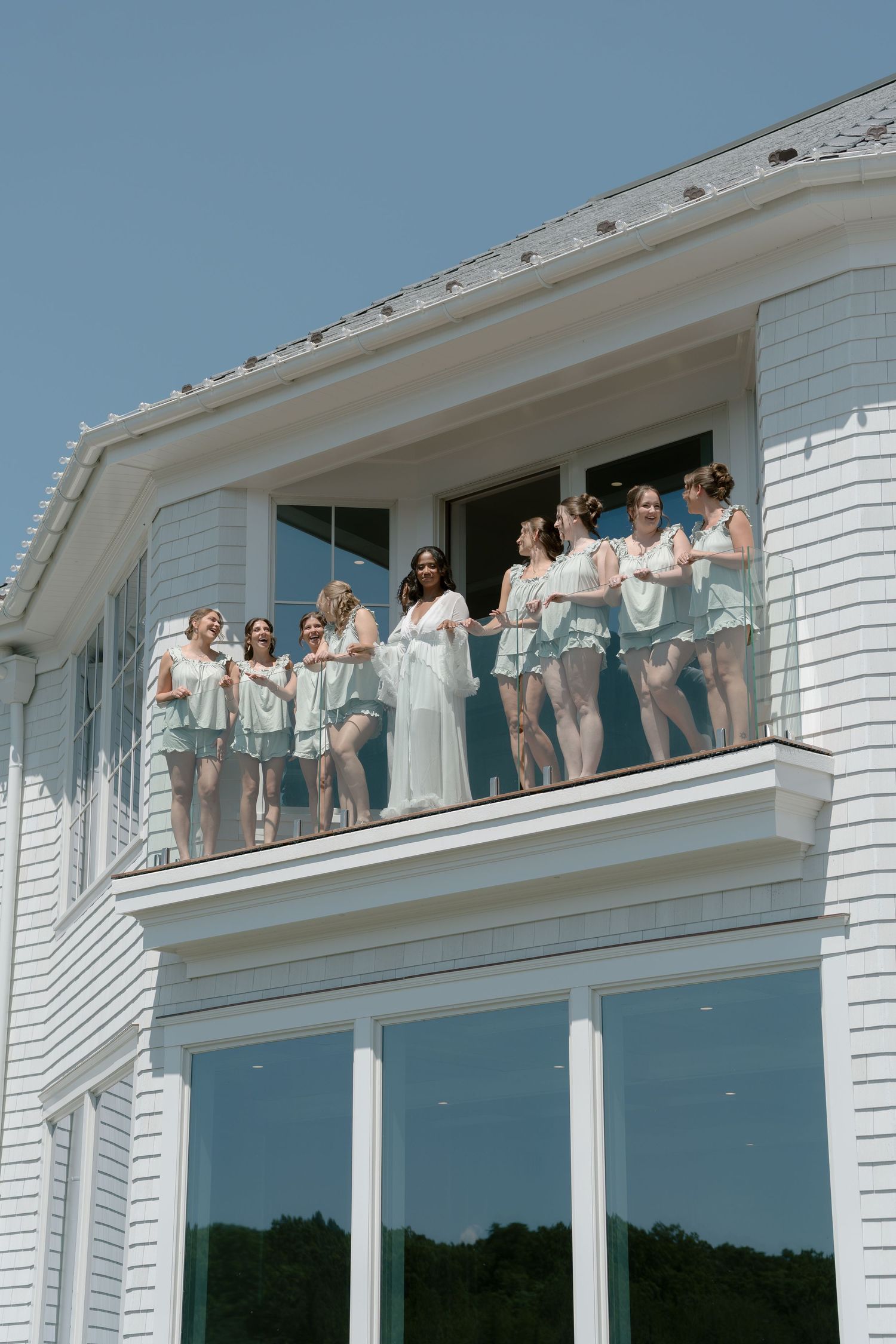 Bridesmaids in matching mint pajamas stand together on a white balcony under a clear blue sky.