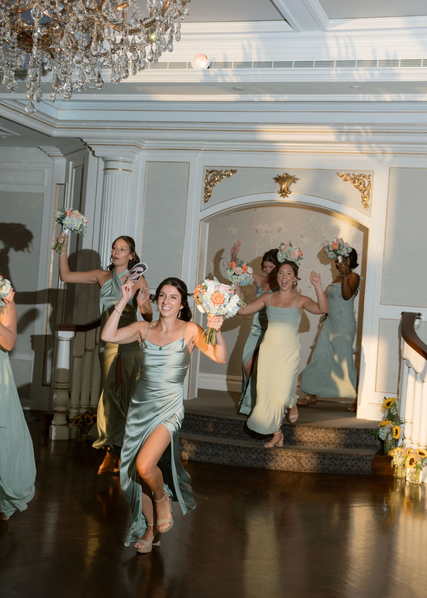 Bridesmaids in sage green satin dresses joyfully entering the wedding reception holding bouquets.