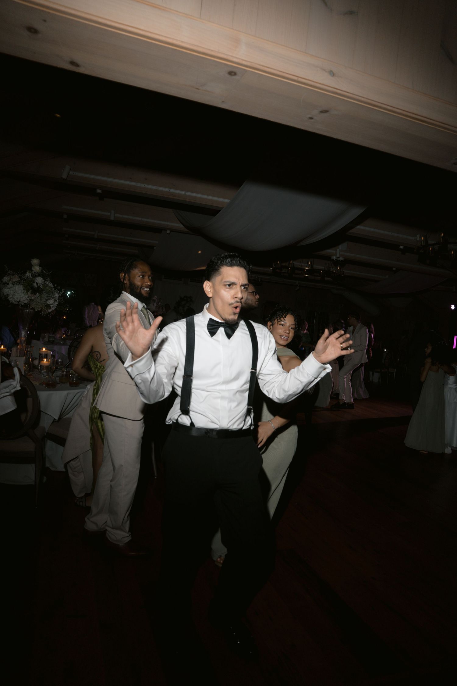 Man in suspenders and bow tie dancing energetically at a formal wedding reception.