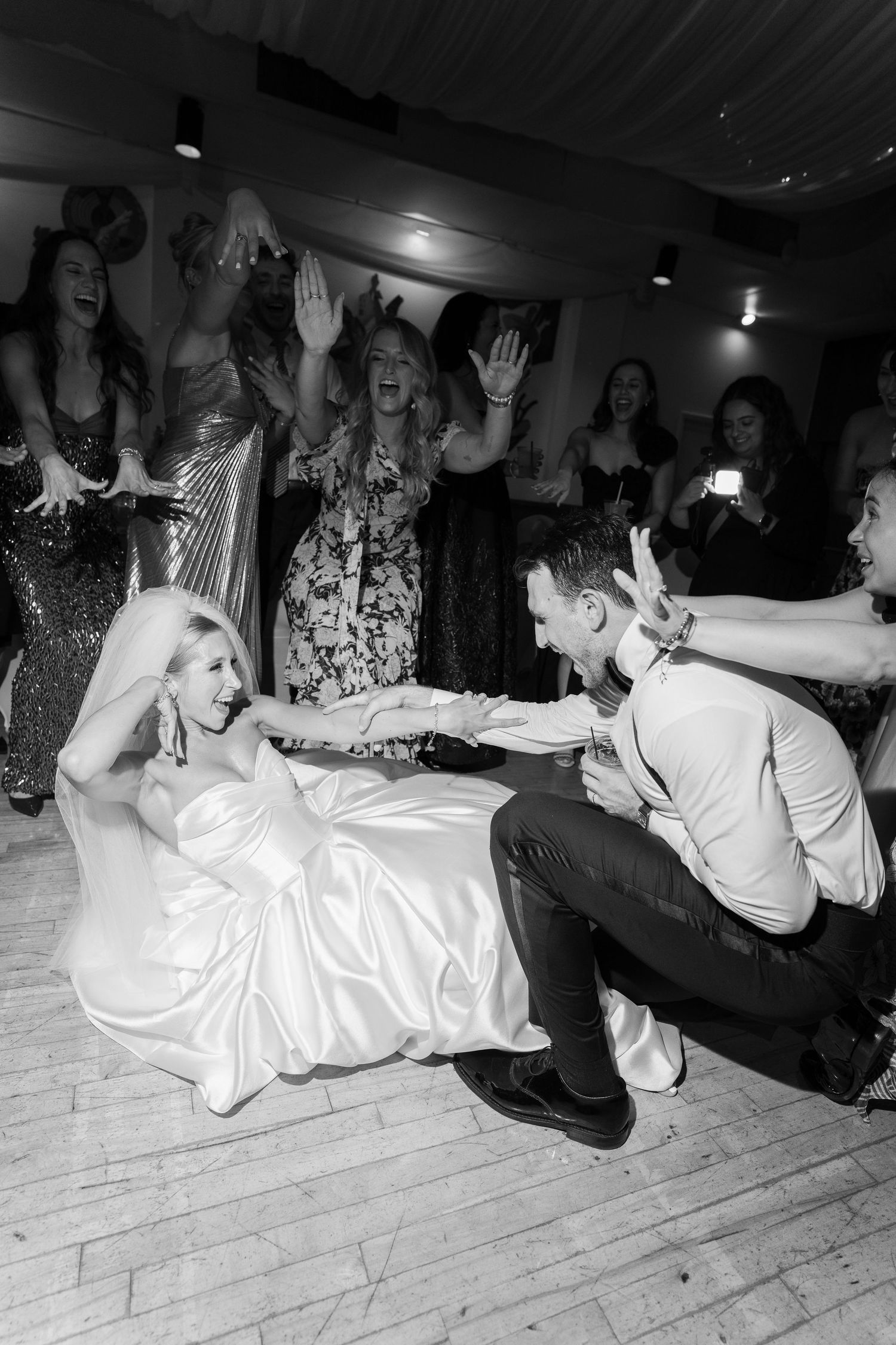 Bride and groom sit on dance floor laughing as guests cheer around them at wedding reception.