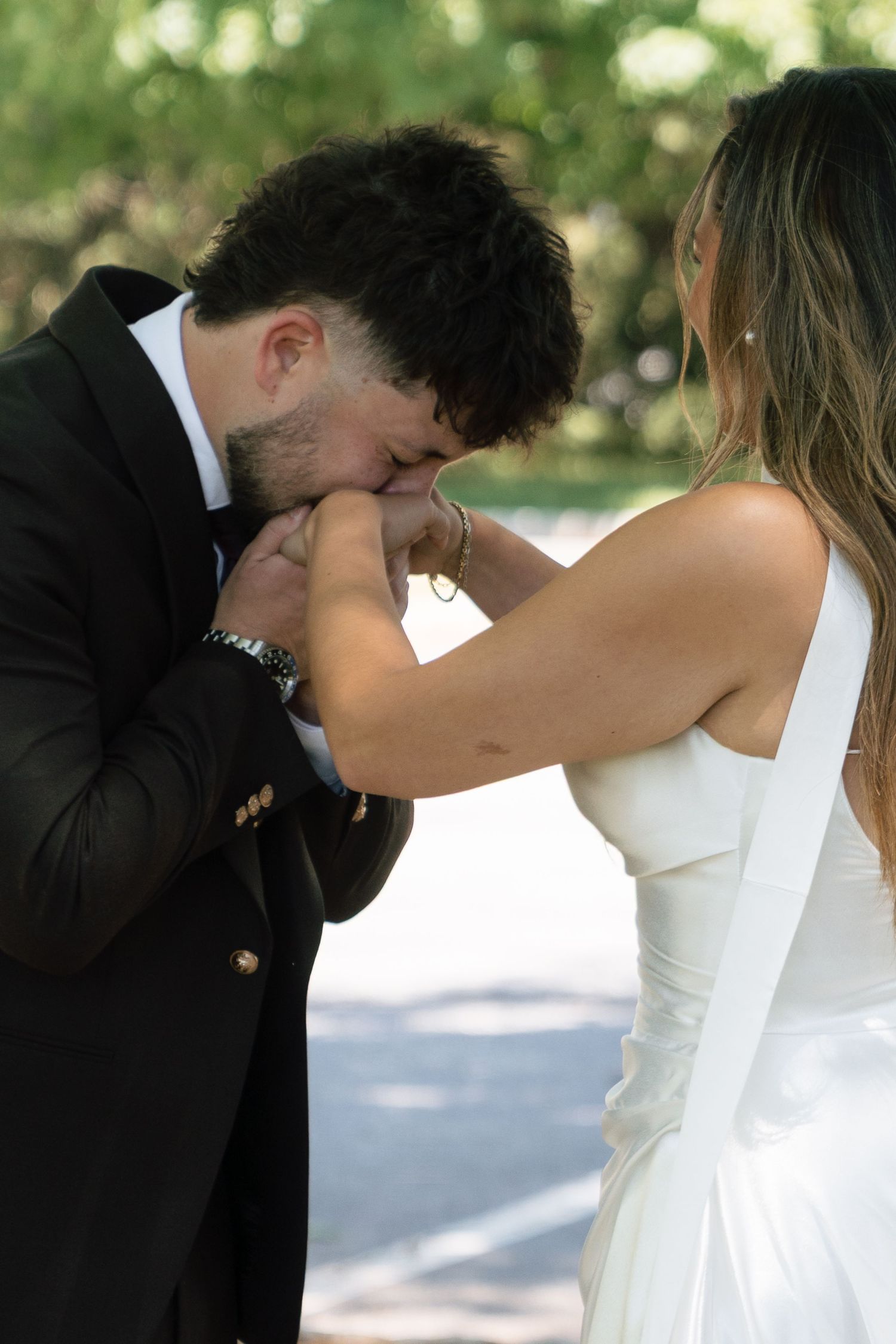Groom in black tuxedo kissing bride's hand tenderly during wedding first look.