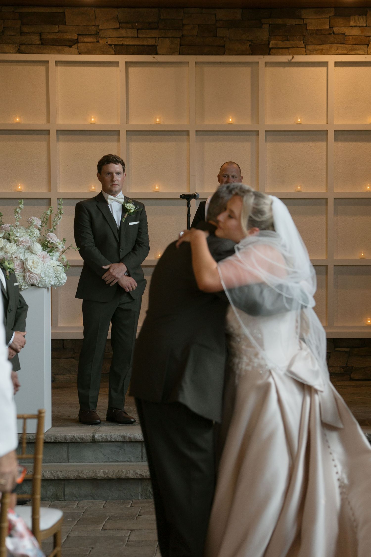 Groom watches bride walk down the aisle at elegant indoor wedding ceremony with candlelit backdrop.