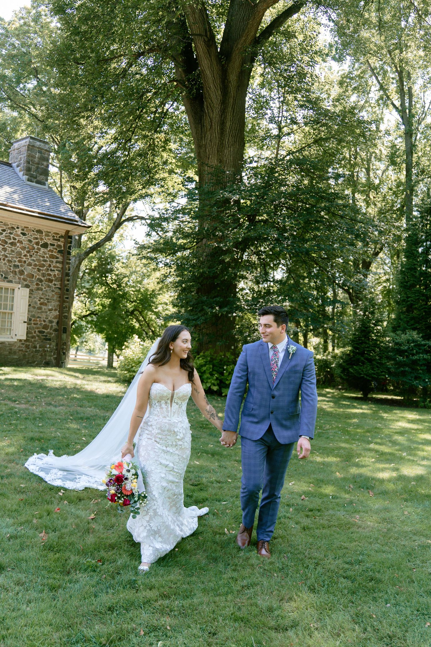 Newlyweds walk hand in hand through a scenic garden with stone buildings in the background.