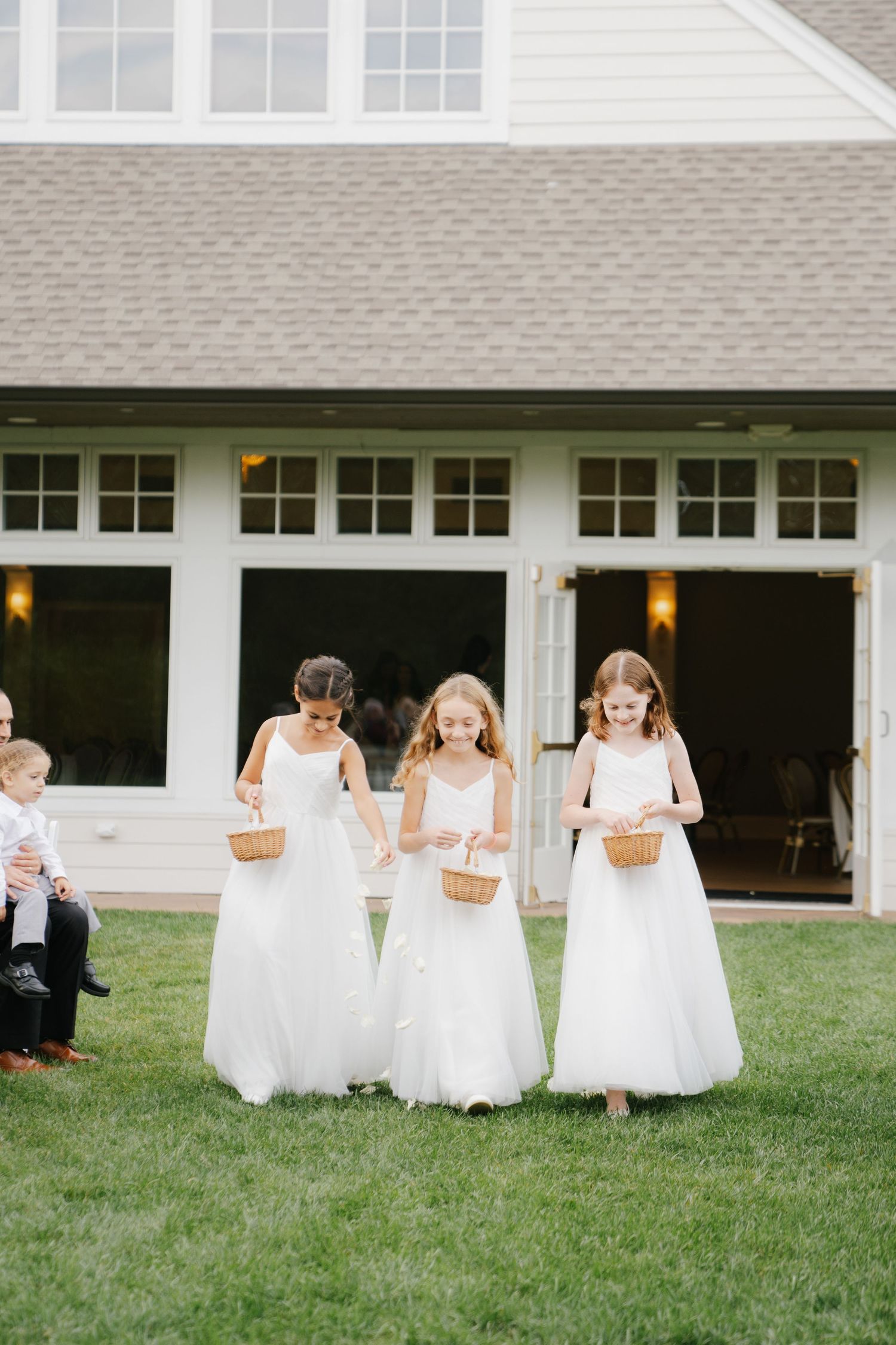A row of flower girls in white dresses run and play on a grassy lawn outside a white building.