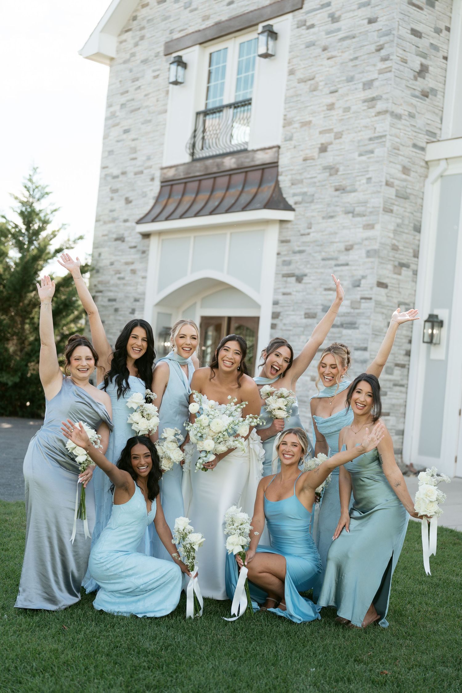 Bride and bridesmaids in blue dresses celebrate joyfully outside a stone mansion.