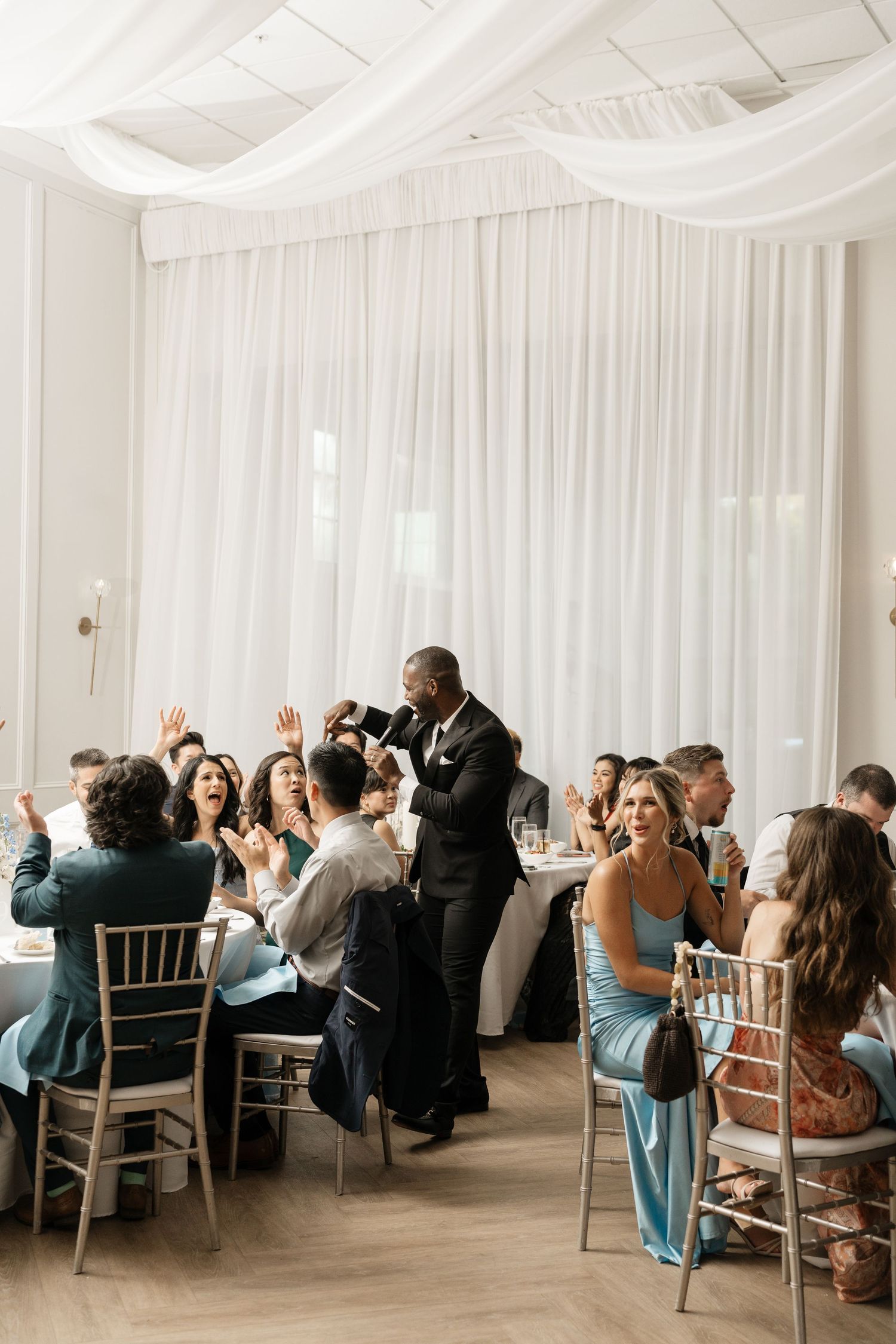 Wedding guests celebrate and applaud during lively reception in elegant white draped ballroom.