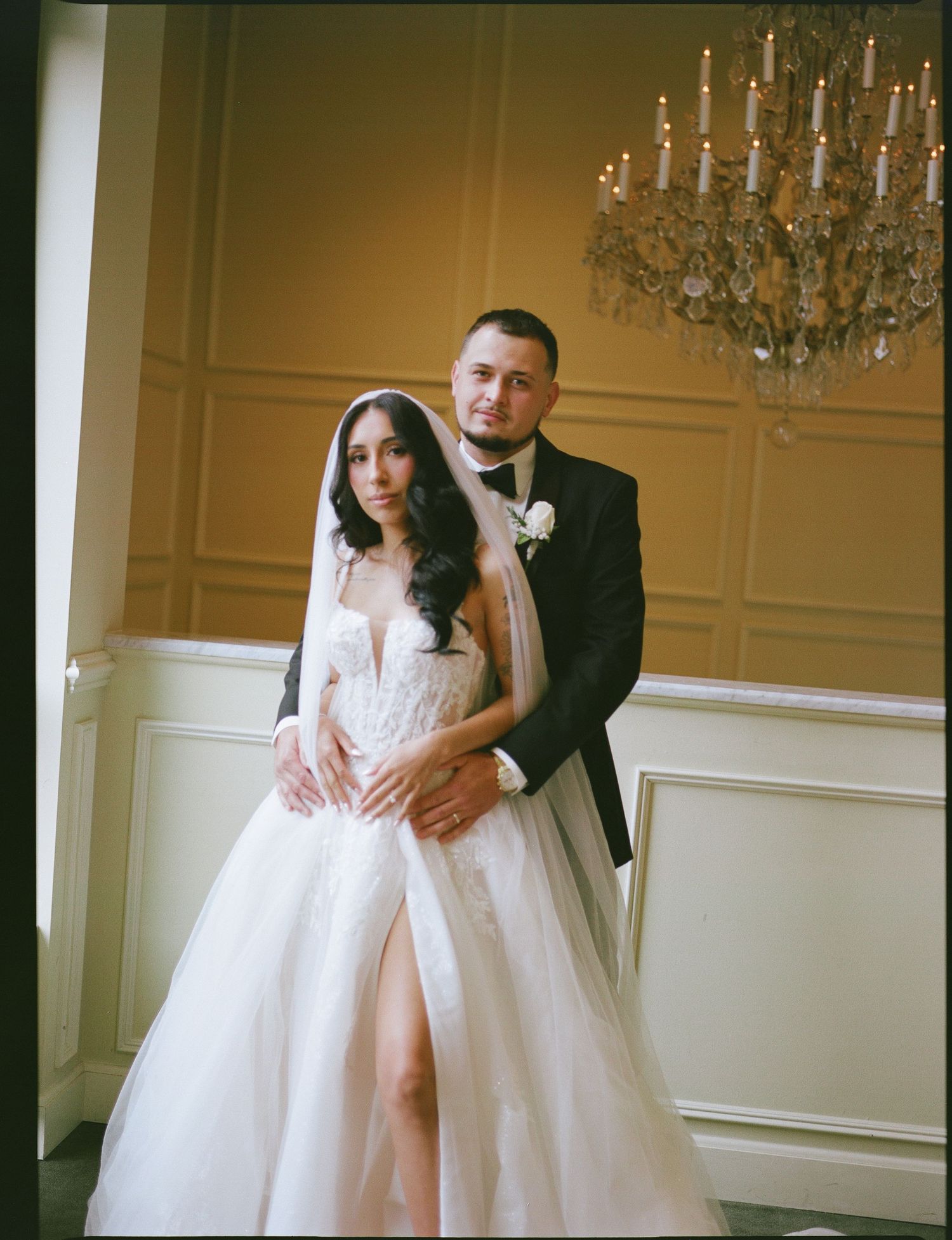 Elegant bride in lace ballgown with veil and groom in tuxedo posing together under chandelier in ornate wedding venue.