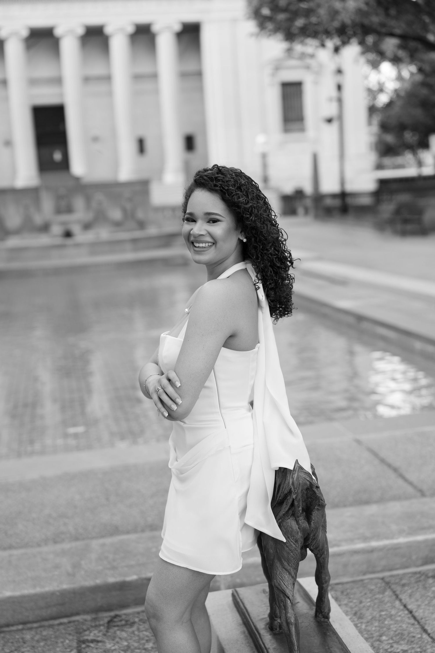 Black and white portrait of smiling woman in white dress near reflective fountain with classical building.
