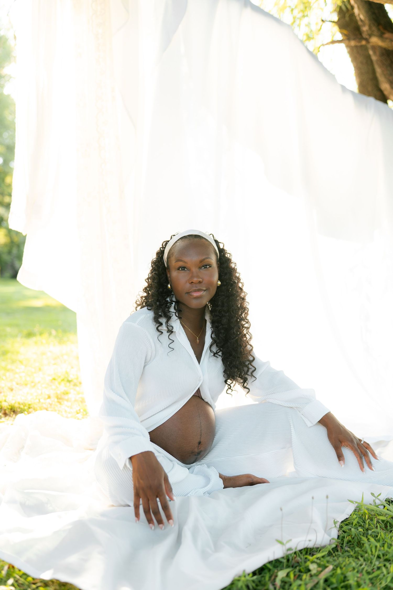 Pregnant Black woman in white outfit sits gracefully on grass outdoors, cradling her baby bump with flowing white drapes behind her.