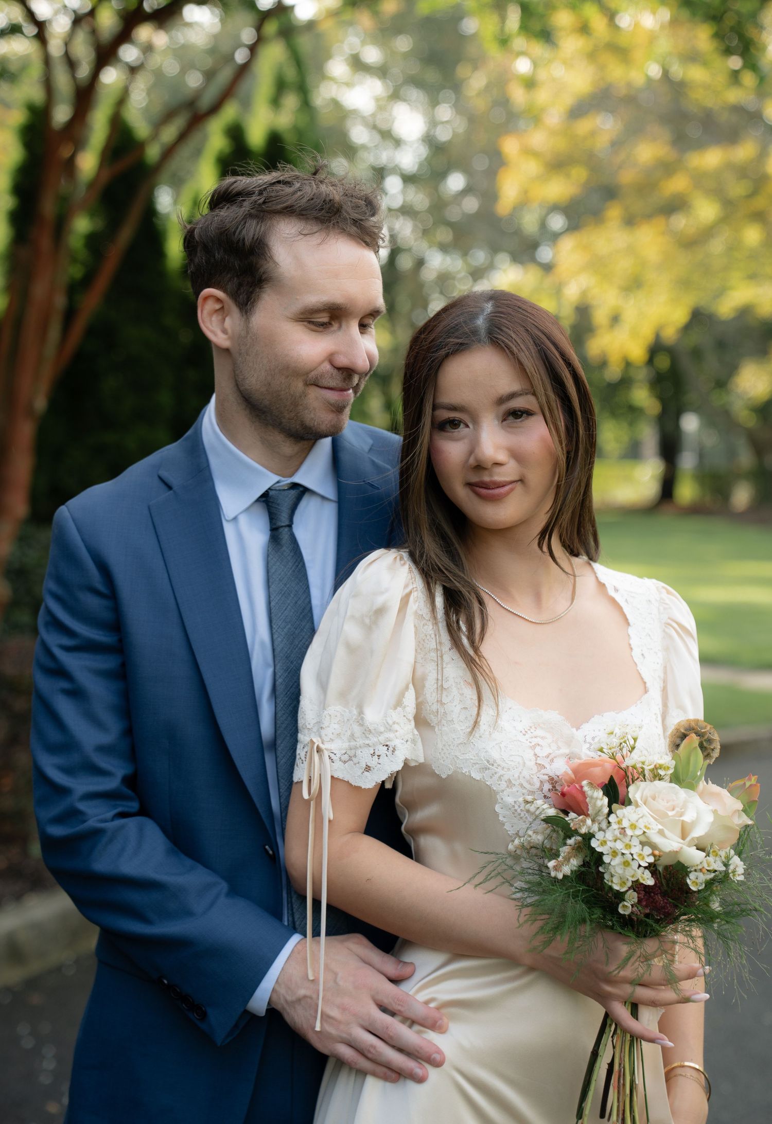 A couple poses together outdoors with autumn trees and natural lighting in the background.