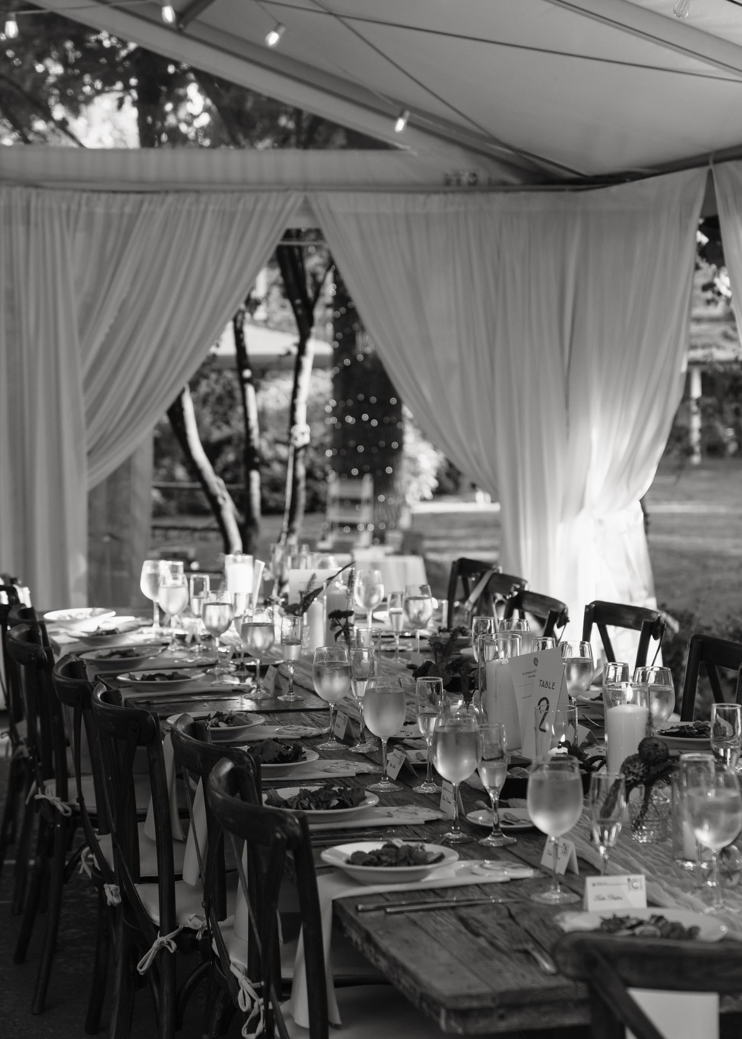Elegant wedding reception tables set under a white tent with rustic wooden chairs and draped linens.