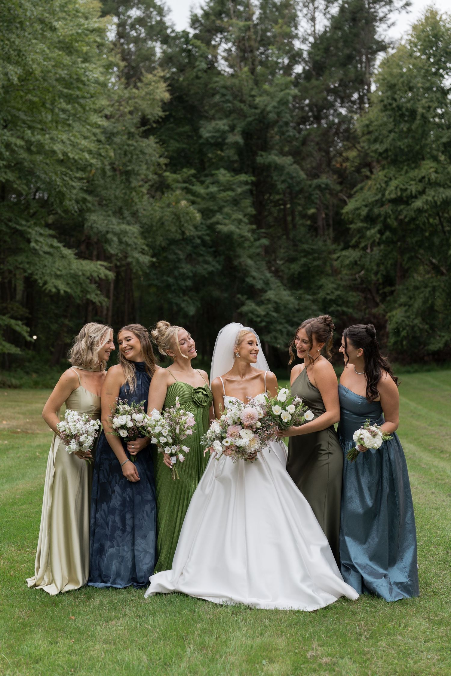 Bride and six bridesmaids in mismatched green and blue gowns laugh together on a grassy lawn.