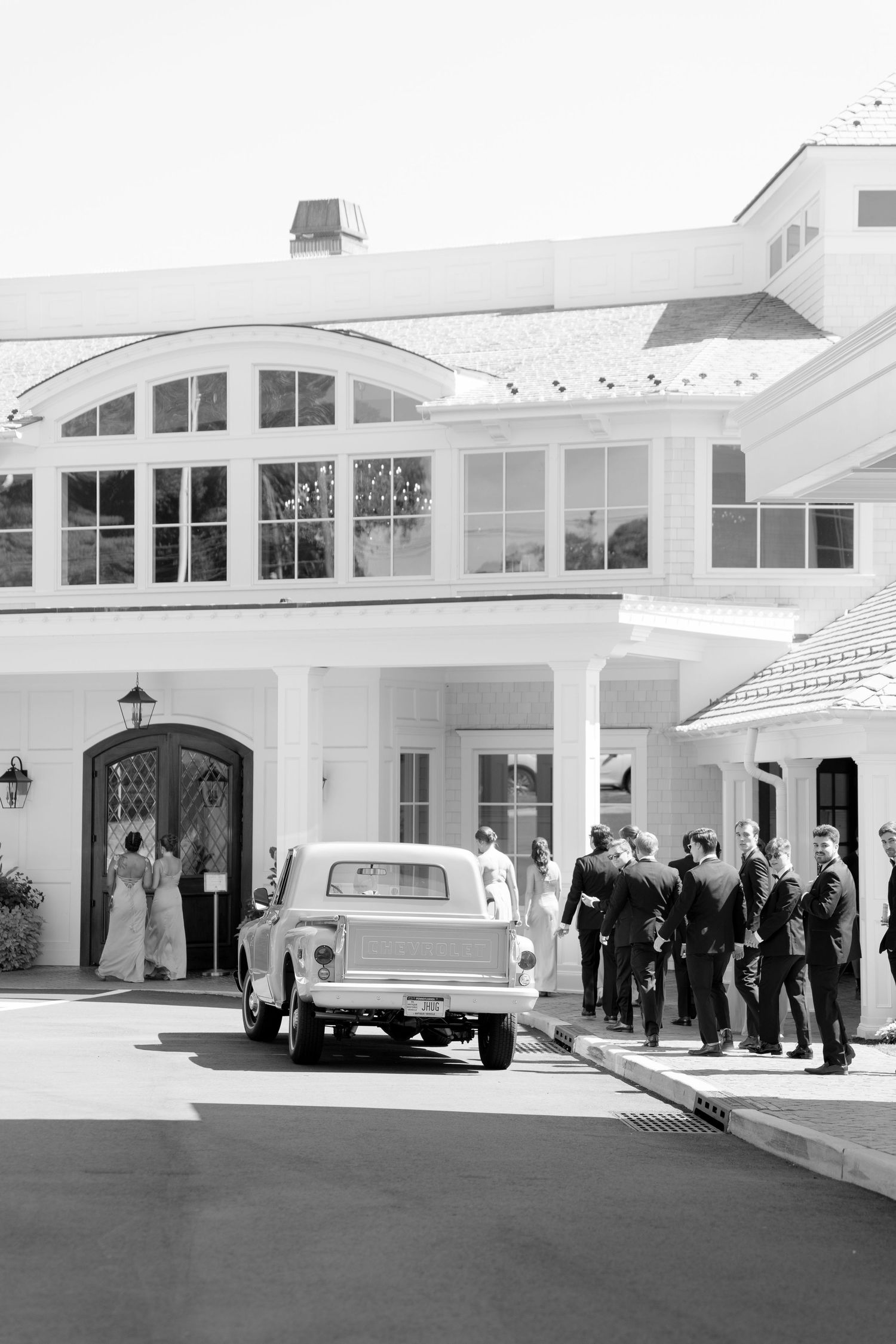 Black and white photo of wedding guests arriving at an elegant white venue with a vintage light-colored truck parked outside.