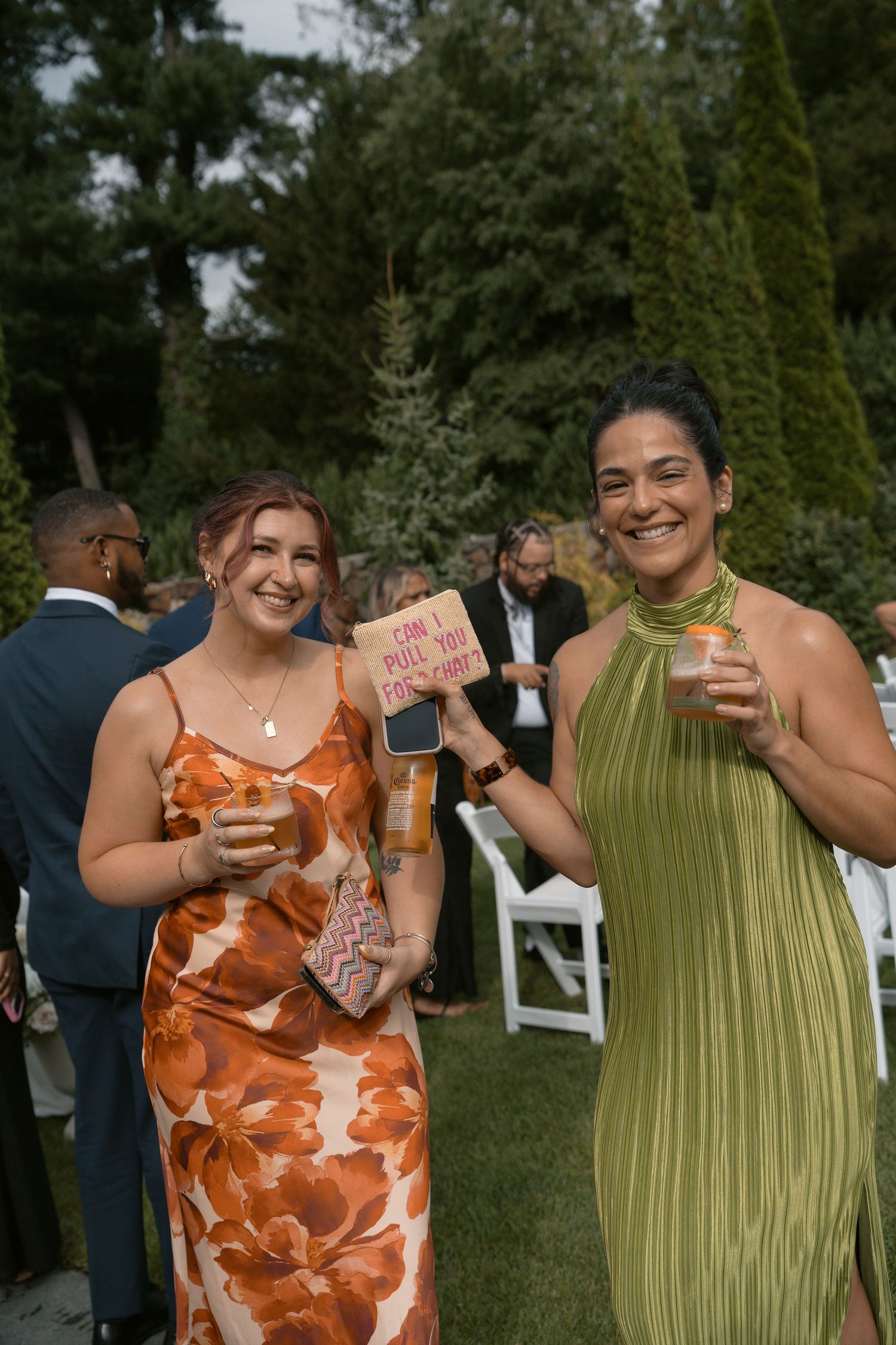 Two smiling women in elegant dresses holding drinks at an outdoor wedding reception surrounded by guests.