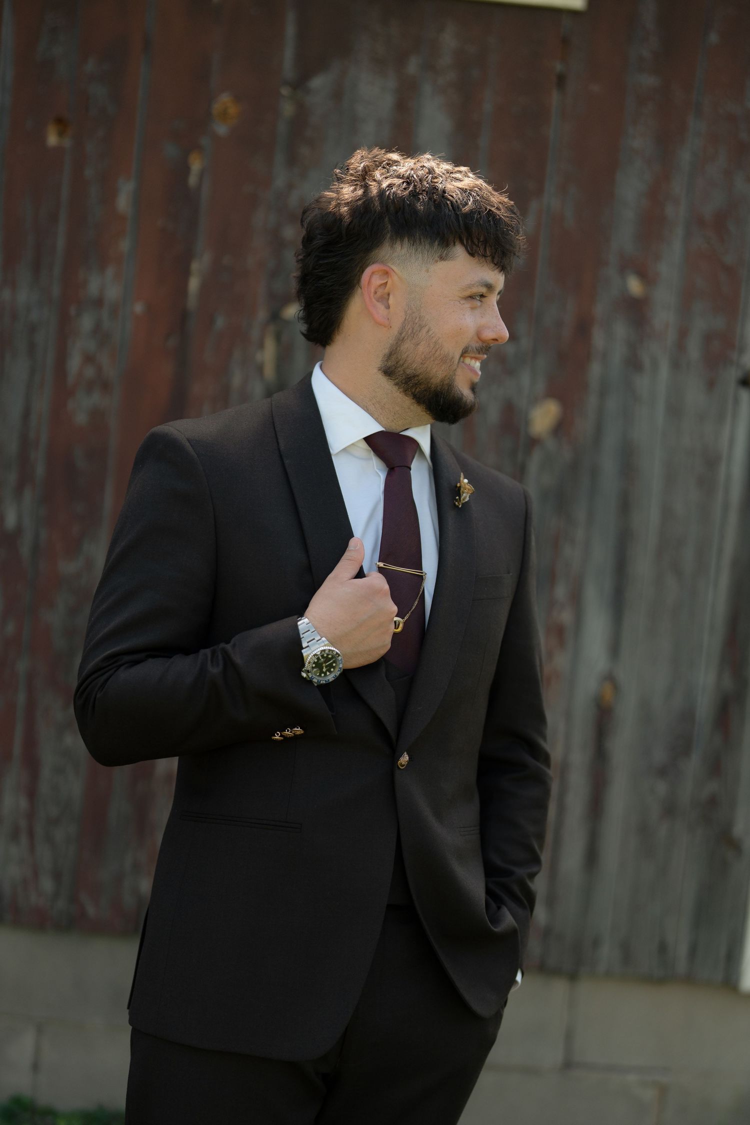 Stylish groom in dark suit with burgundy tie and boutonniere posing confidently outdoors near rustic barn wall.