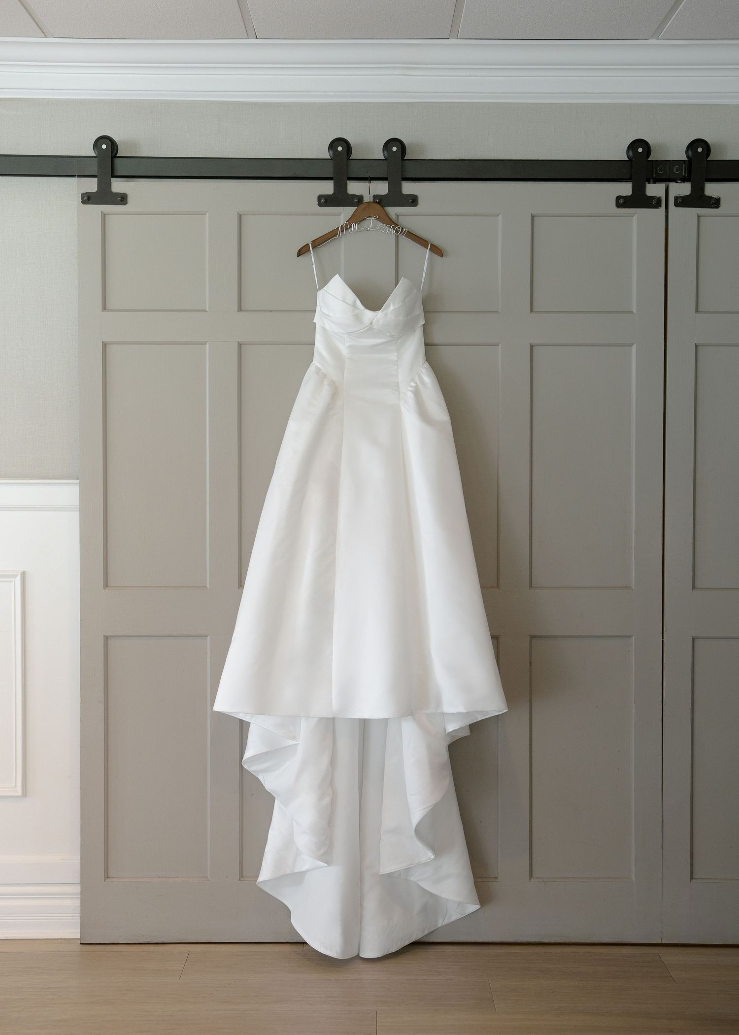 Elegant white strapless A-line wedding gown with high-low skirt hanging on wooden hanger against gray barn doors.