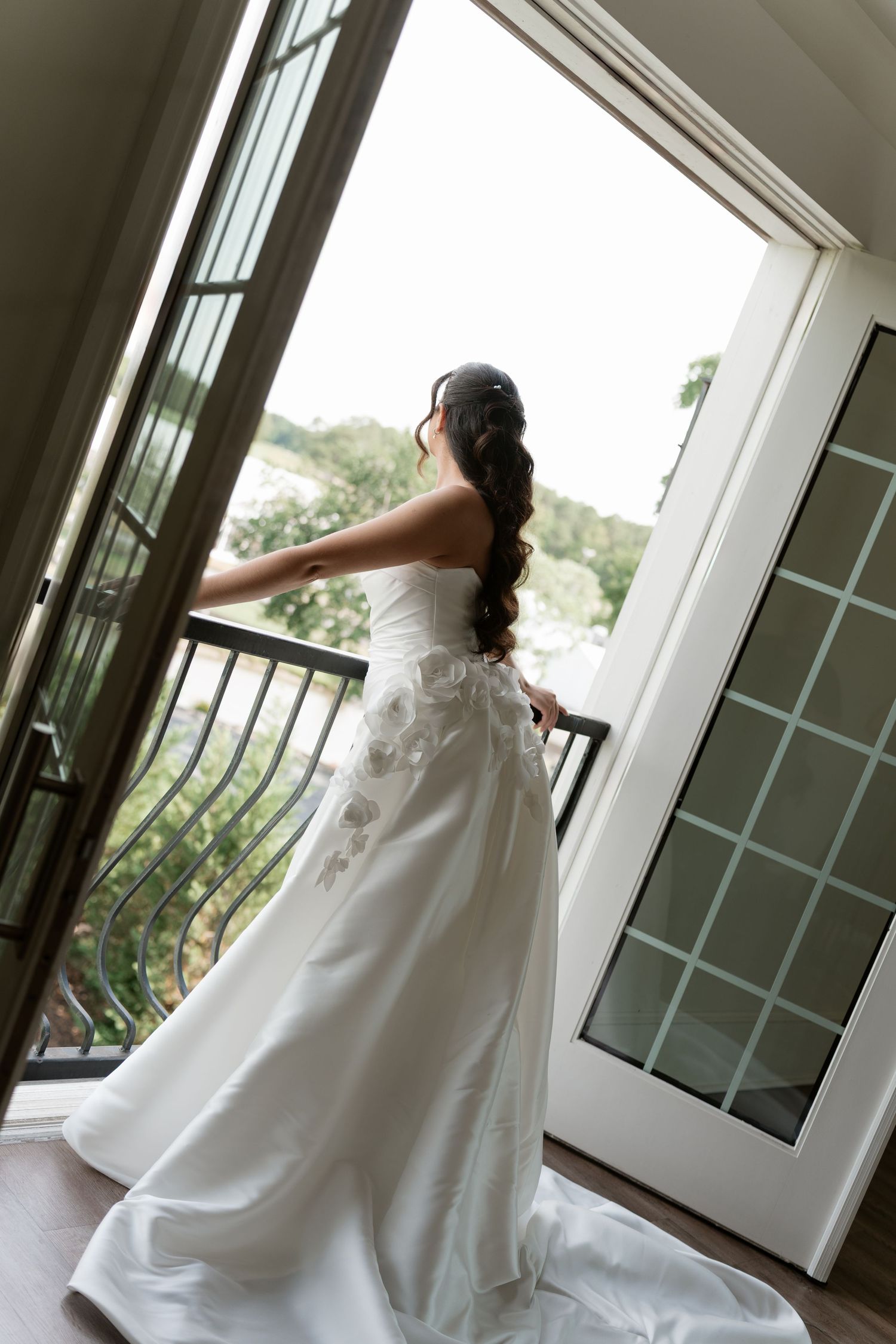 Bride in white strapless wedding gown with floral details stands at open balcony doors, long dark hair cascading down her back.