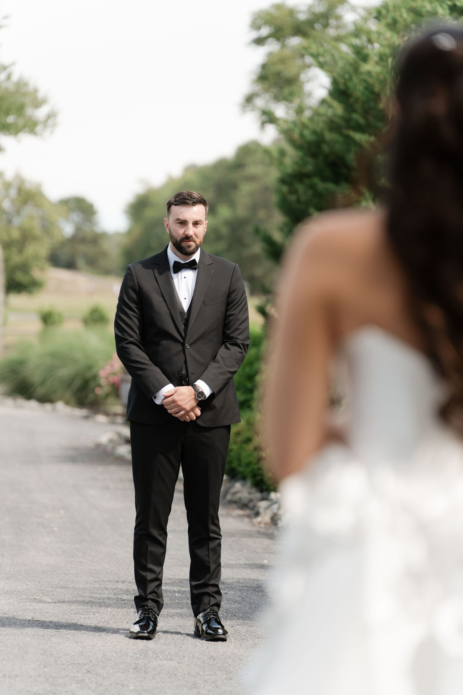 Groom in black tuxedo with bow tie watches bride approach during outdoor first look ceremony.