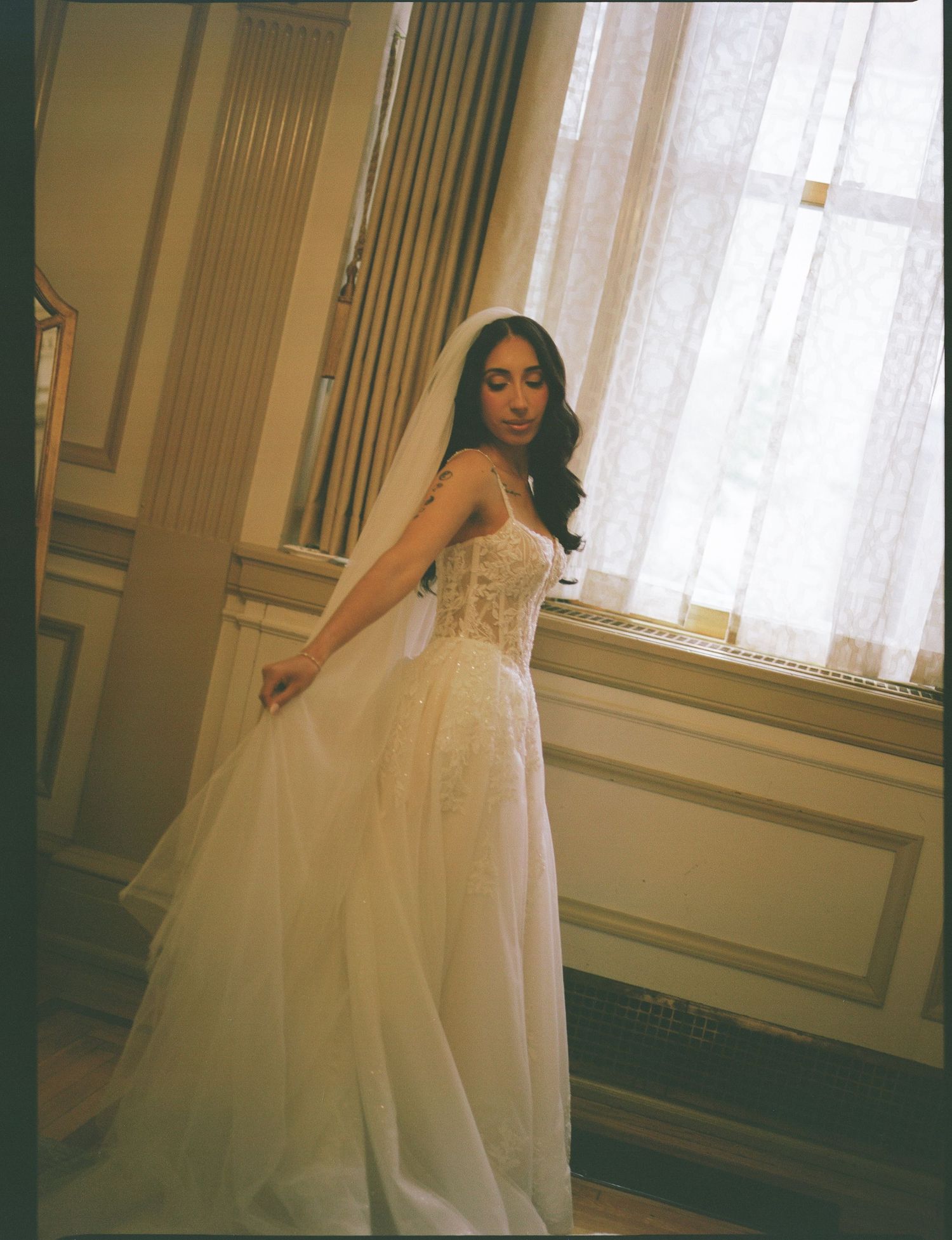Bride in a stunning white lace corset wedding gown with tulle skirt and veil, posing elegantly by a window.