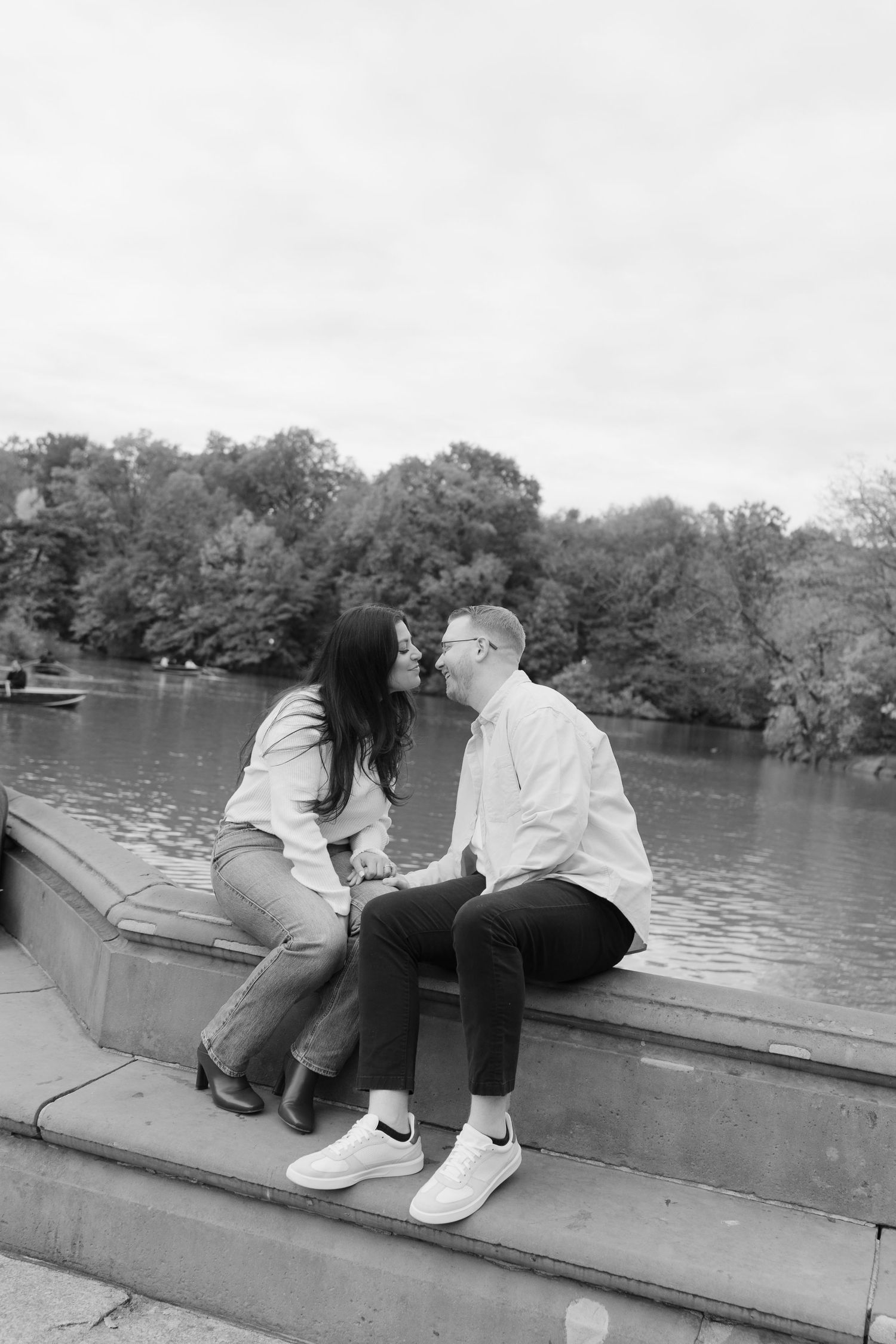 A couple sits together on stone steps overlooking a serene lake surrounded by autumn trees in black and white.