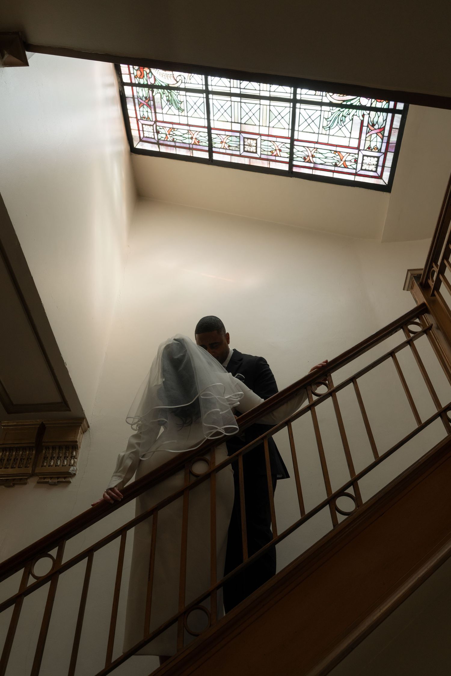 A wedding couple descends an elegant staircase with stained glass windows overhead.