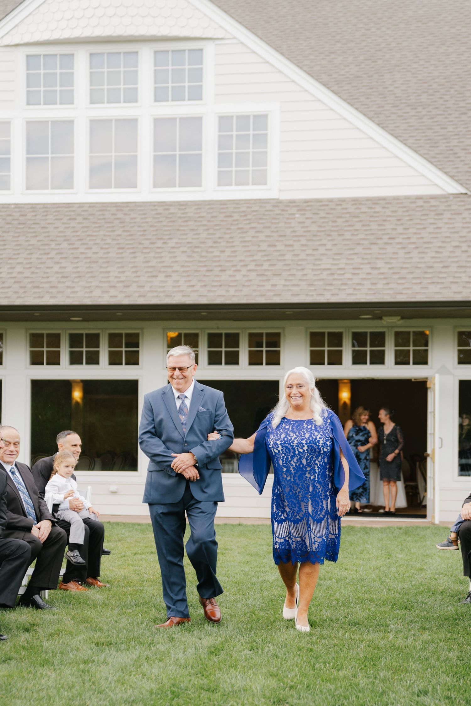 Wedding party members run playfully across a grassy lawn in front of a white house with garage doors.