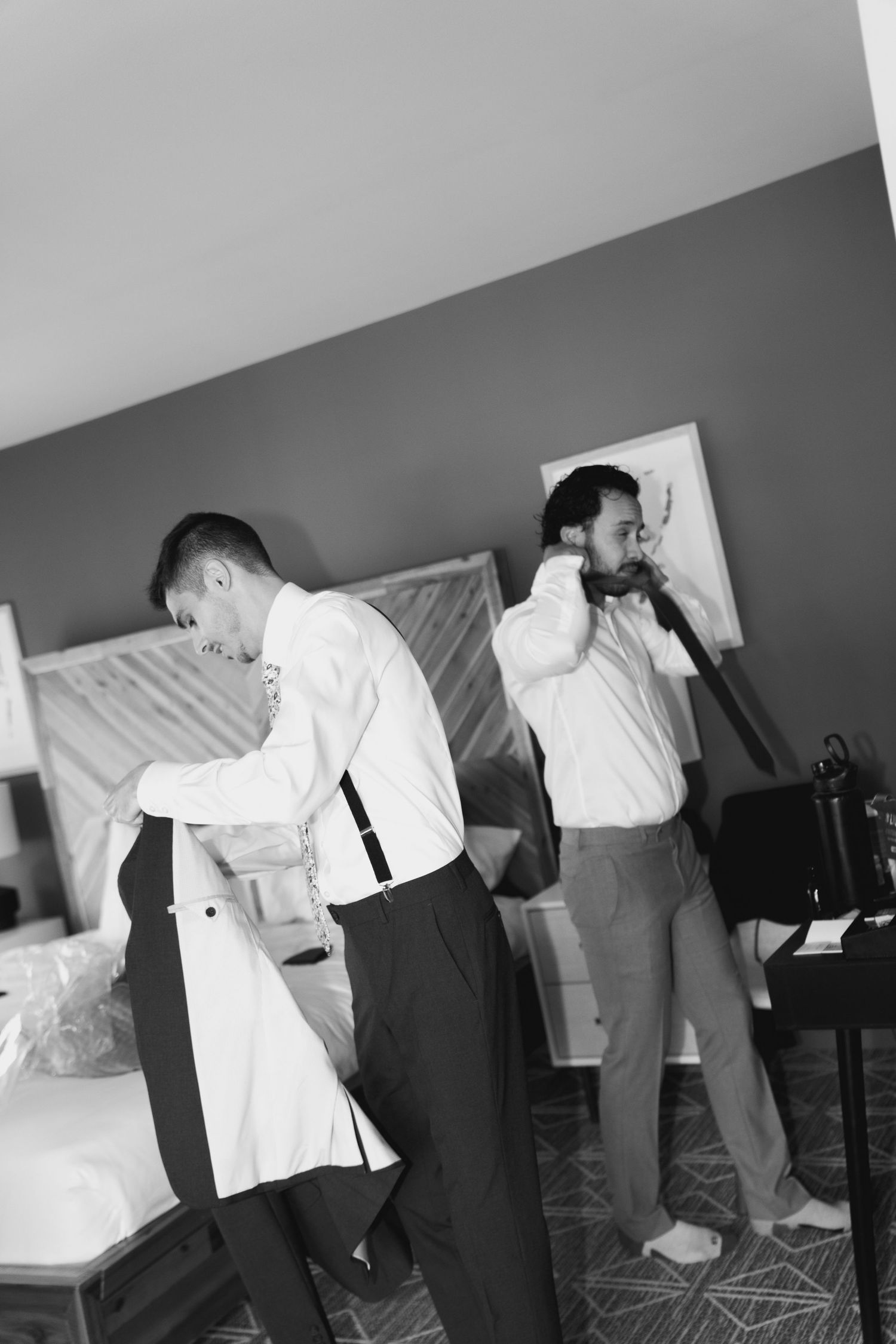 Black and white photo of two men in white shirts and dress pants getting ready in a bedroom.