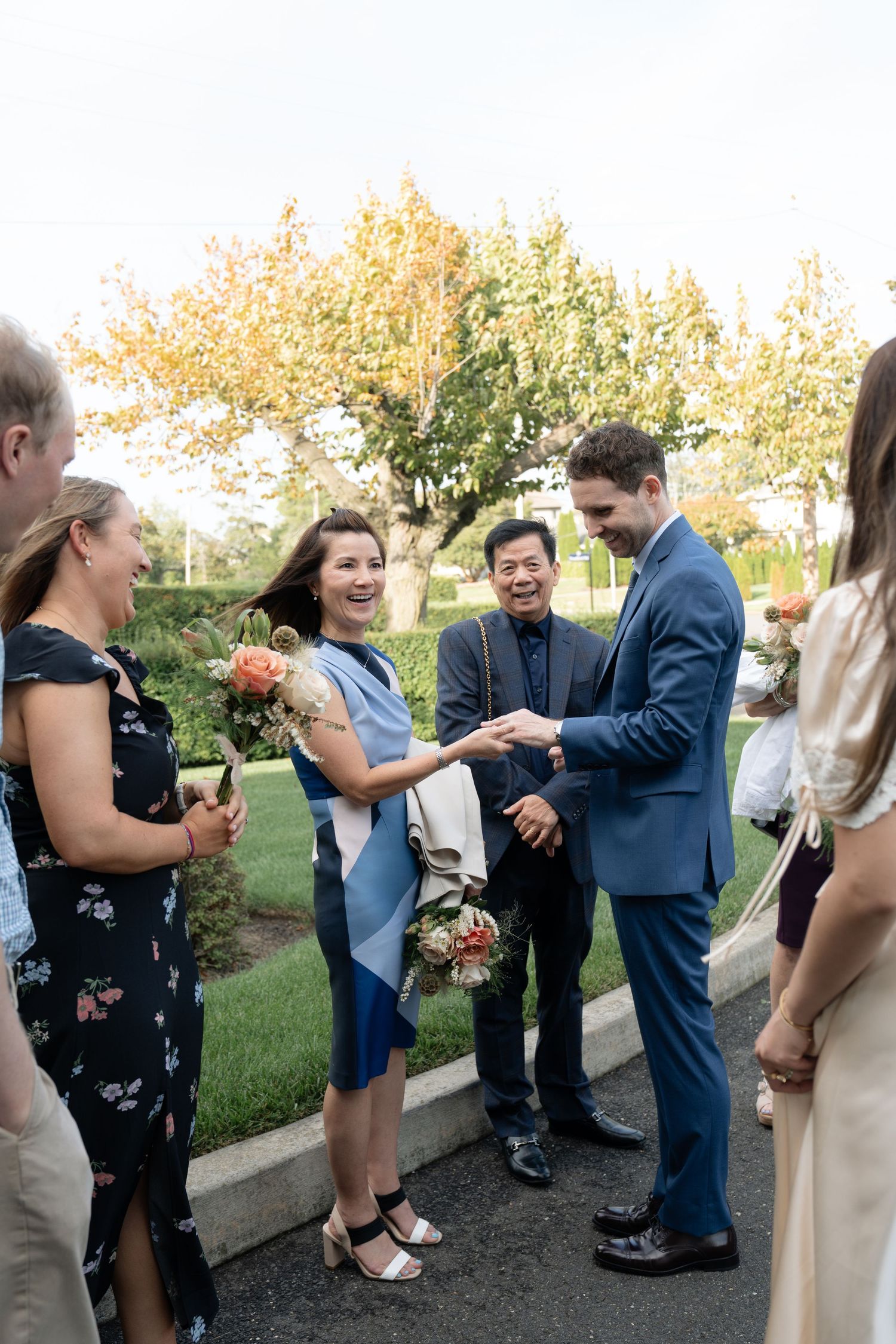 Wedding guests mingle outdoors on a sunny day holding floral bouquets.