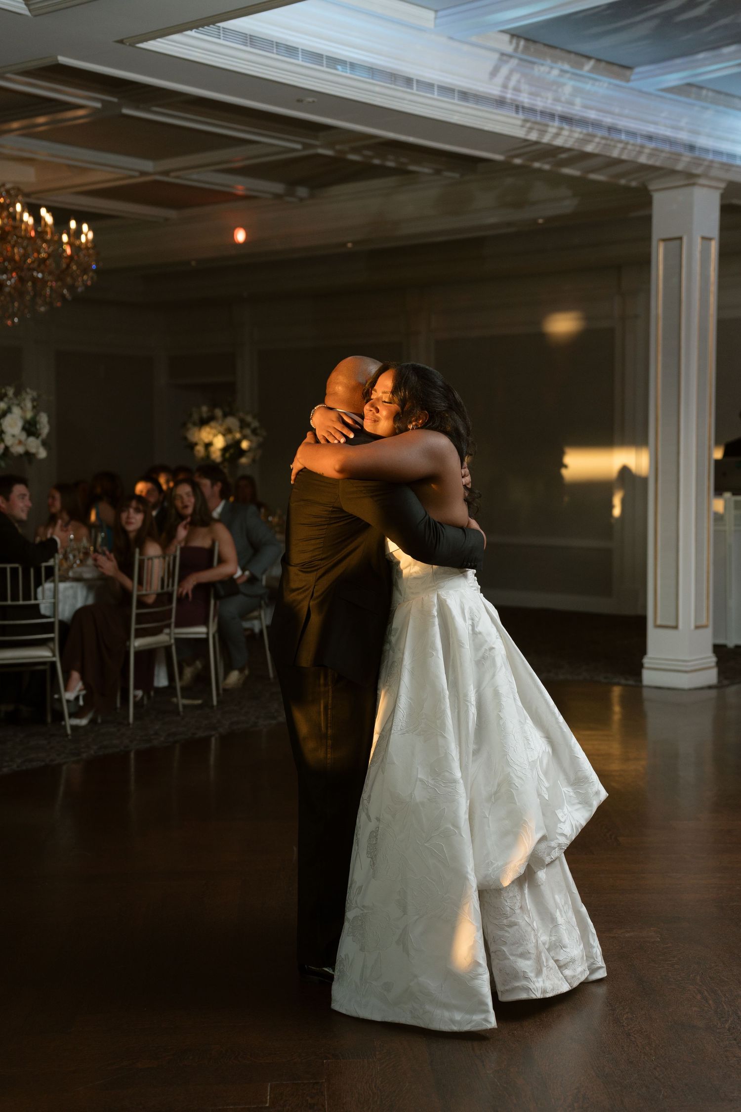 Bride and father share an emotional first dance hug at elegant wedding reception with guests seated in background.