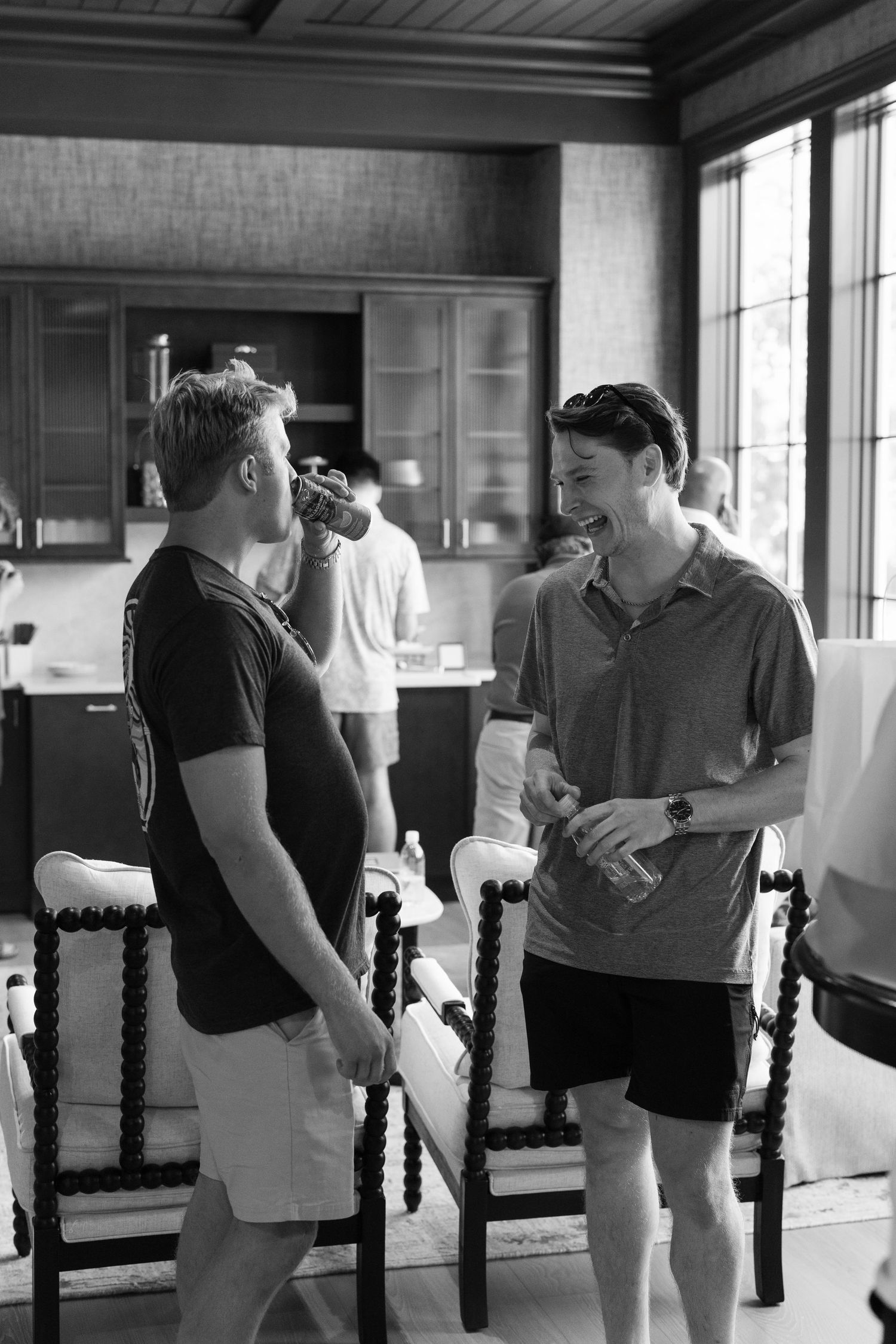 Two groomsmen laughing and drinking beverages together in a elegant kitchen during wedding preparations.
