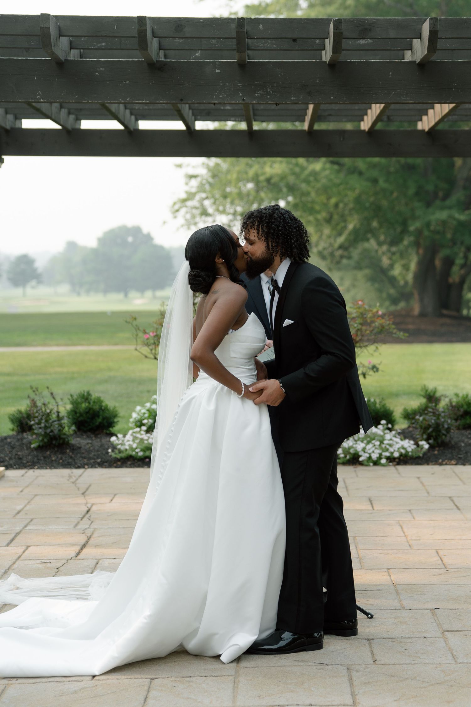 Newlywed couple shares their first kiss as bride and groom under a pergola surrounded by lush greenery.