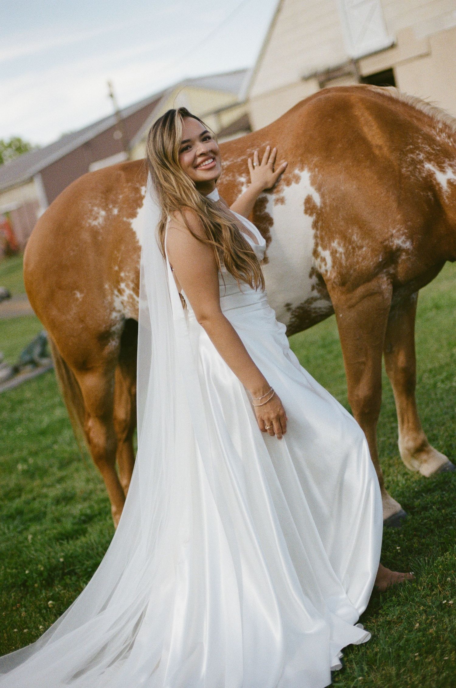 Smiling bride in white wedding gown and veil poses beside a brown and white horse on green grass at a farm.