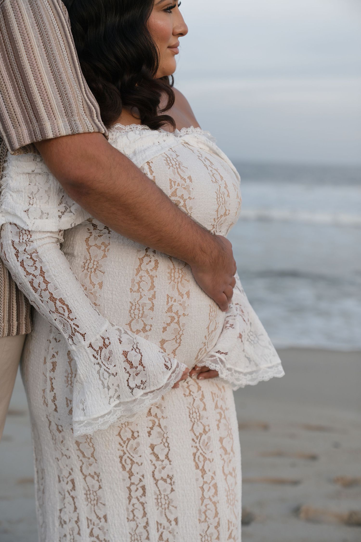 Pregnant woman in white lace off-shoulder gown embraced by partner on beach at dusk.
