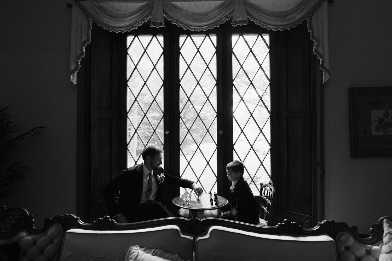 Black and white interior photo of a room with diamond-patterned windows and draped curtains creating a moody atmosphere.