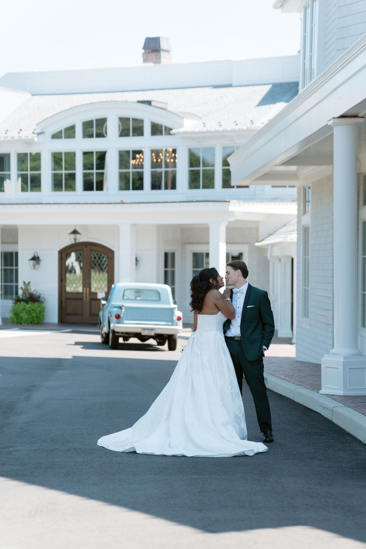 Bride and groom share a romantic moment outside a white colonial wedding venue with a vintage blue car in the background.